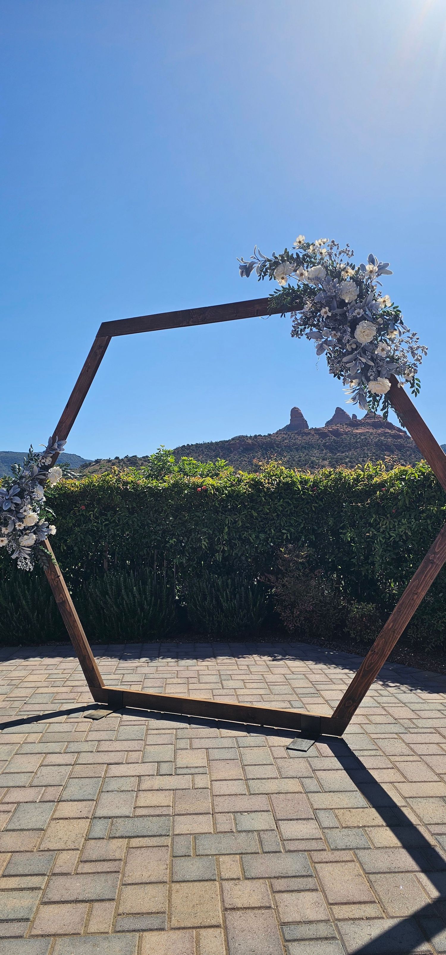 Hexagonal wooden wedding arch adorned with white floral arrangements on a brick patio with mountain views.