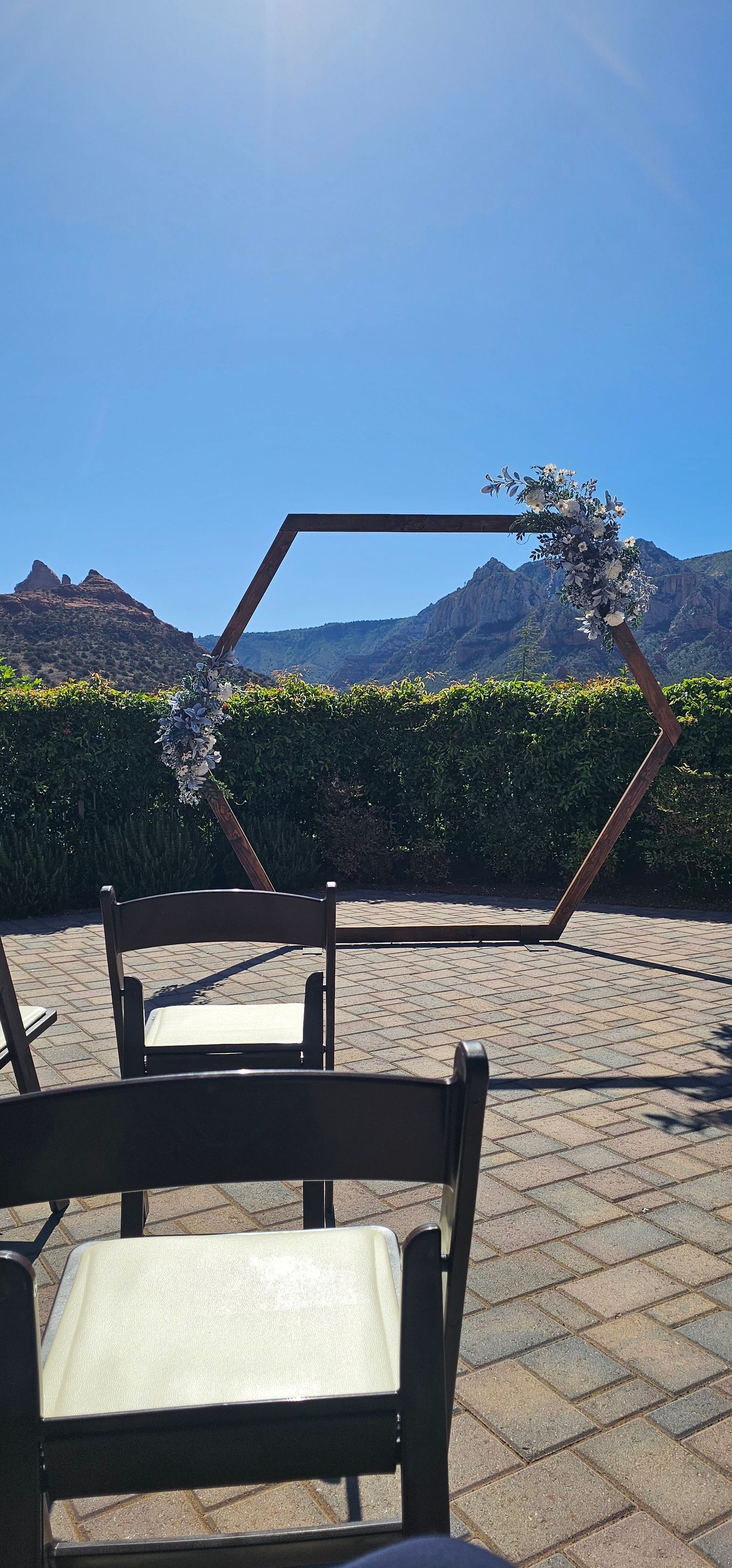 Outdoor wedding ceremony setup with hexagonal wooden arch adorned with flowers, dark chairs on brick patio, and mountain backdrop.