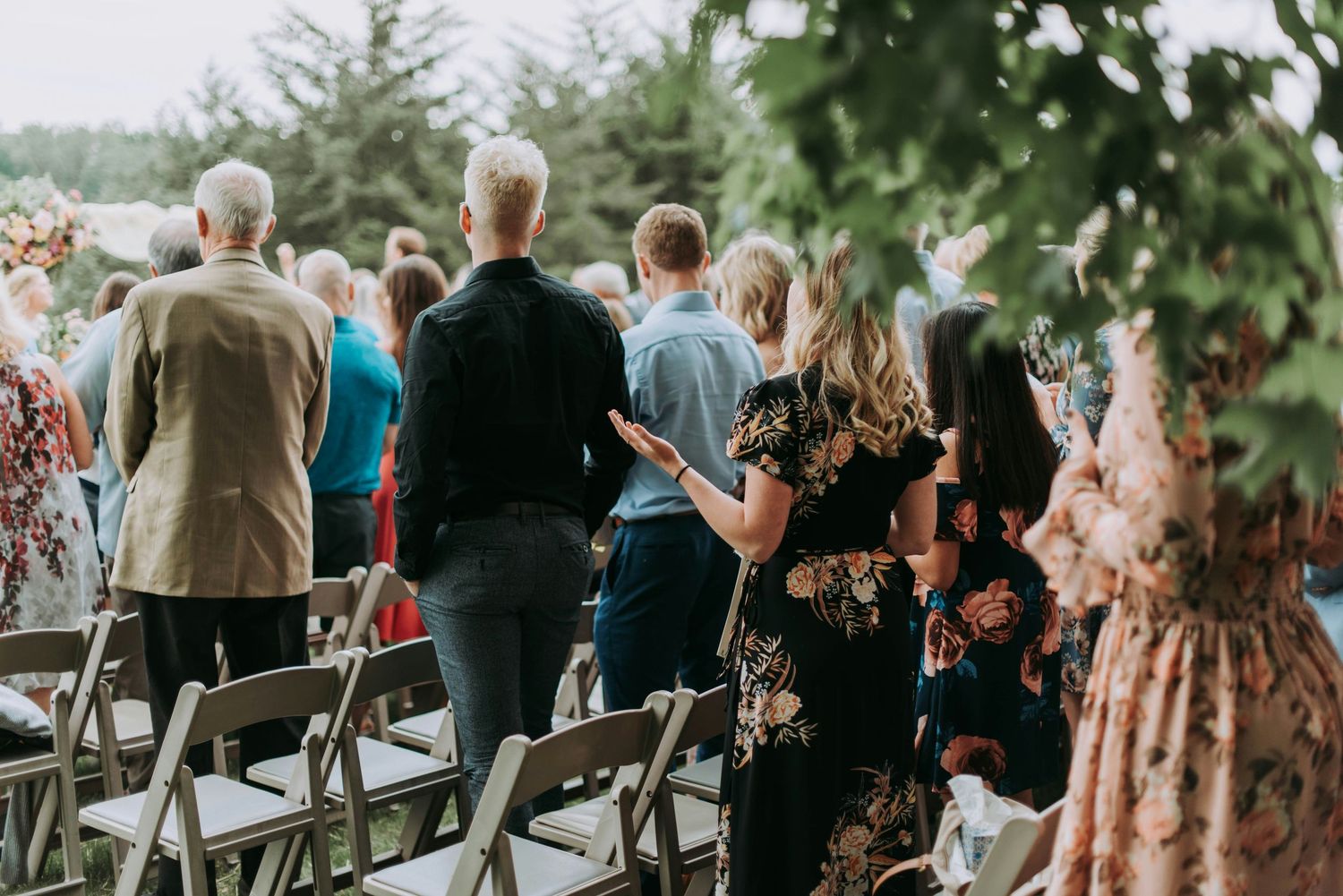 Guests standing and walking between rows of gray folding chairs at an outdoor wedding ceremony surrounded by trees.