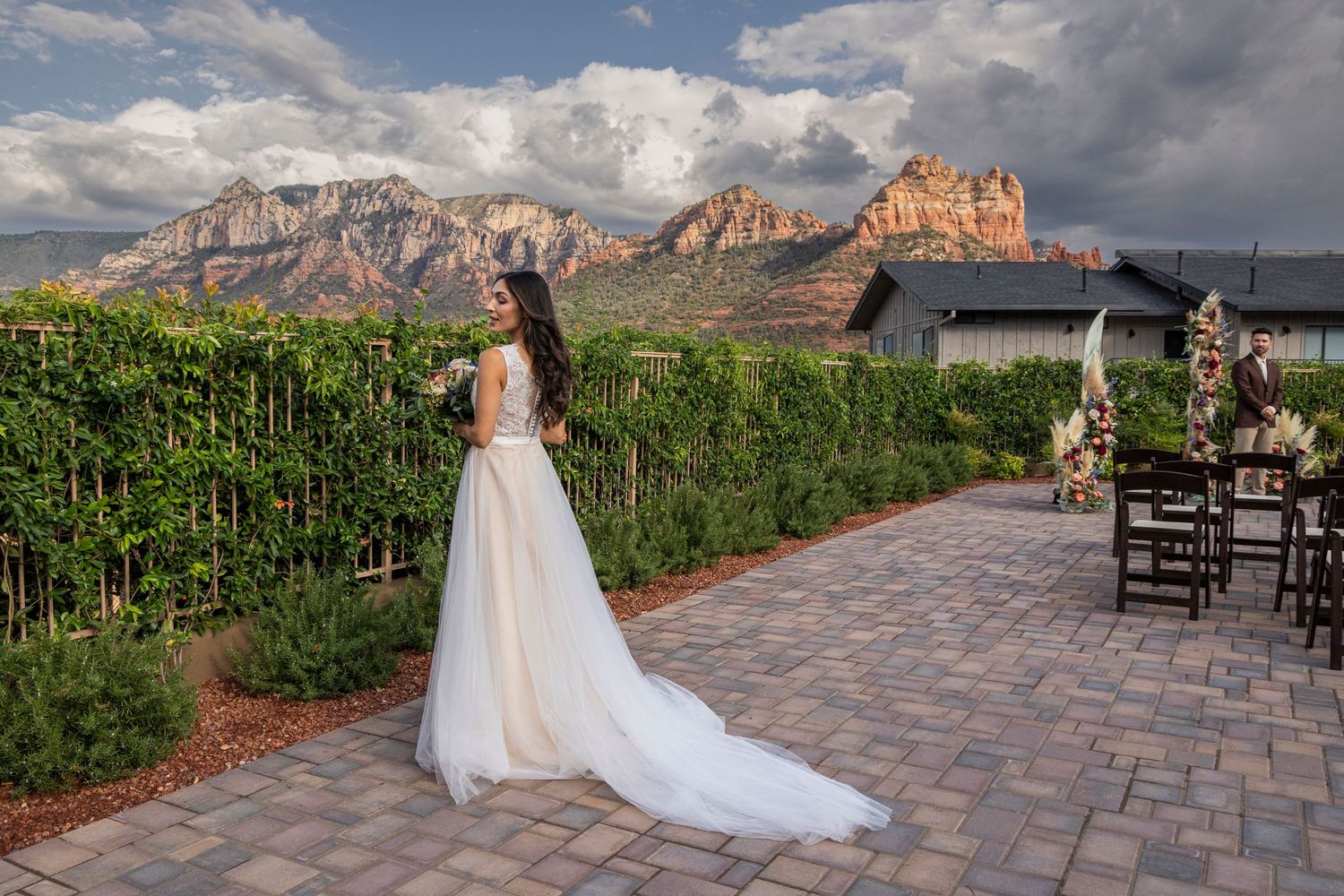 Bride in flowing white gown stands in vineyard with stunning Sedona red rock mountains backdrop on cloudy day.