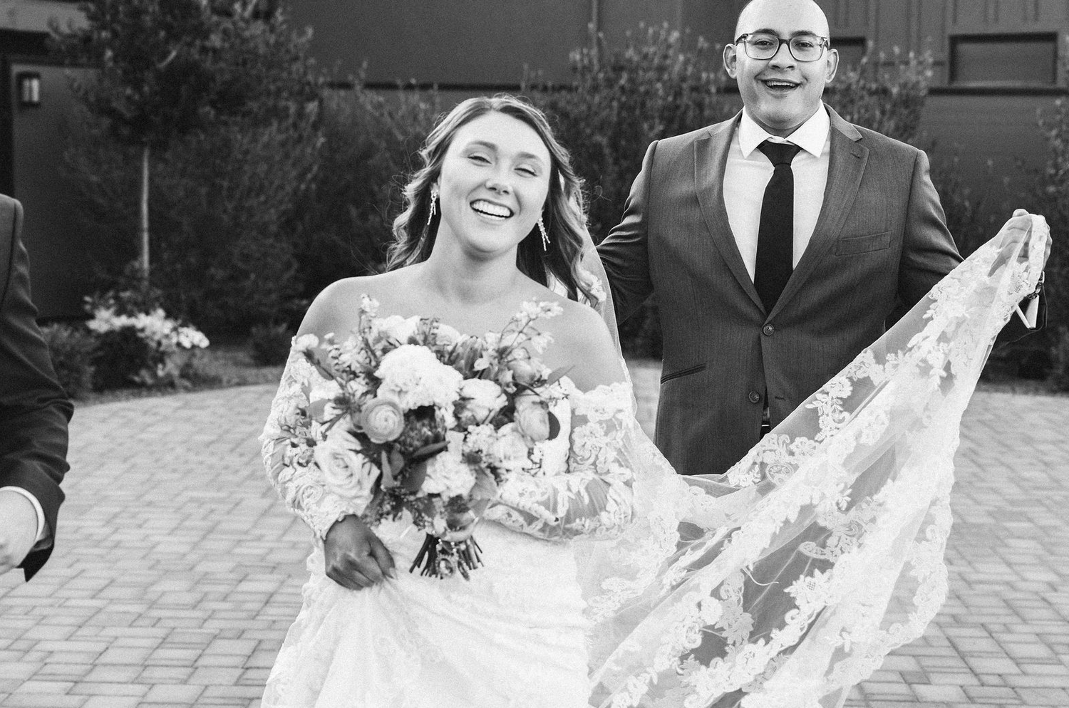 Black and white photo of a smiling bride holding a bouquet with her groom, veil flowing, on their wedding day.