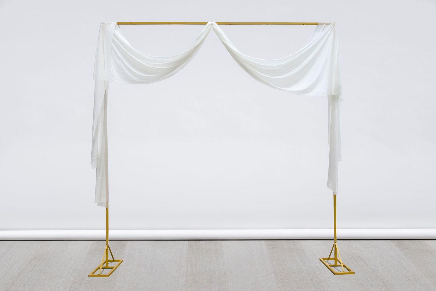 Wedding arch frame with double-draped white fabric and gold metal base stands against white wall.