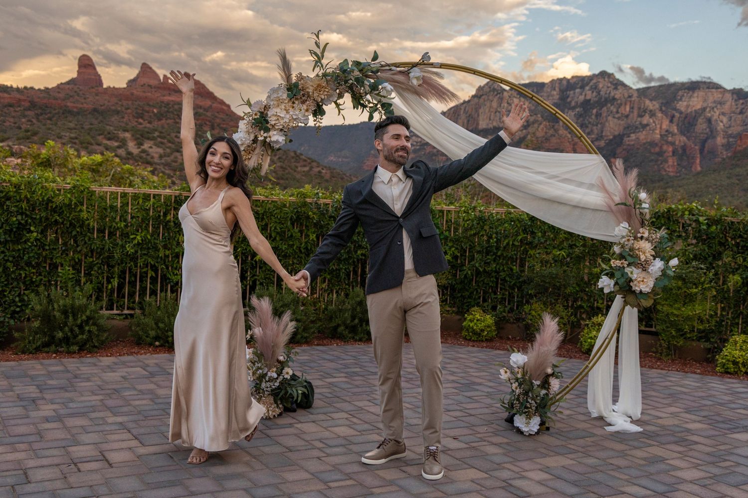 Couple celebrating wedding ceremony outdoors in Sedona, Arizona, with red rock mountains and floral arch backdrop.