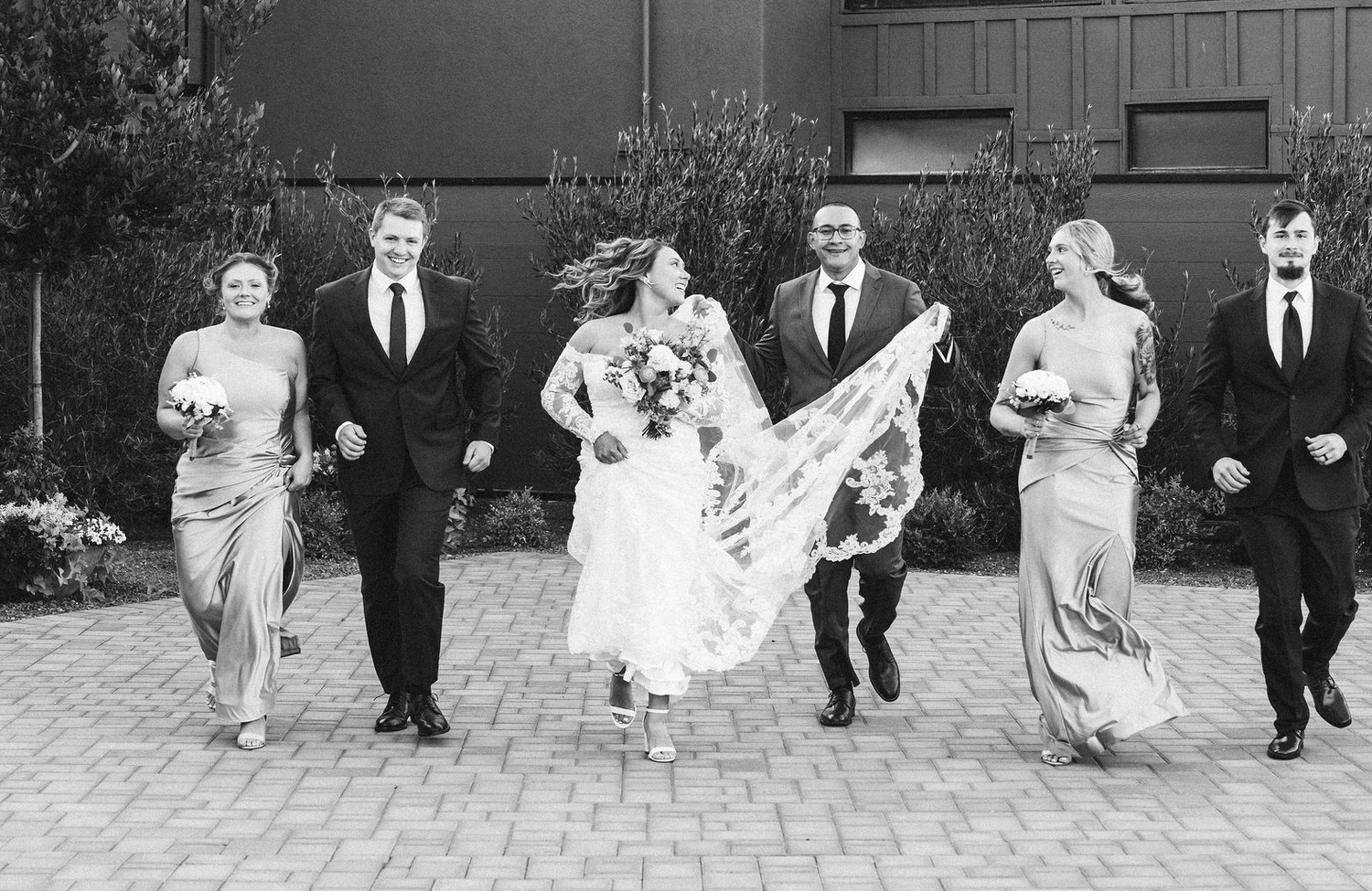 Black and white photo of a joyful wedding party walking together outdoors, bride's veil flowing in the wind.