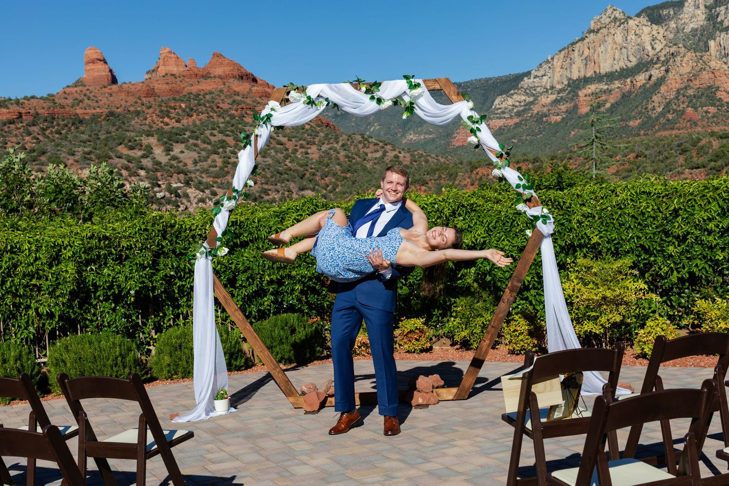 Groom lifts bride under floral arch at outdoor Sedona wedding ceremony with red rock formations backdrop.