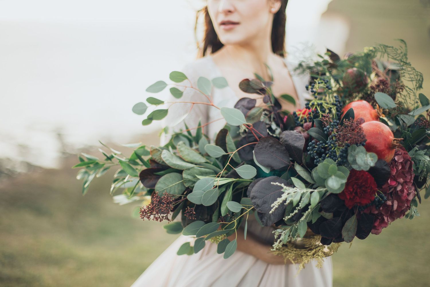 Bride holding a lush oversized bouquet with dark berries, eucalyptus, and deep red flowers outdoors.