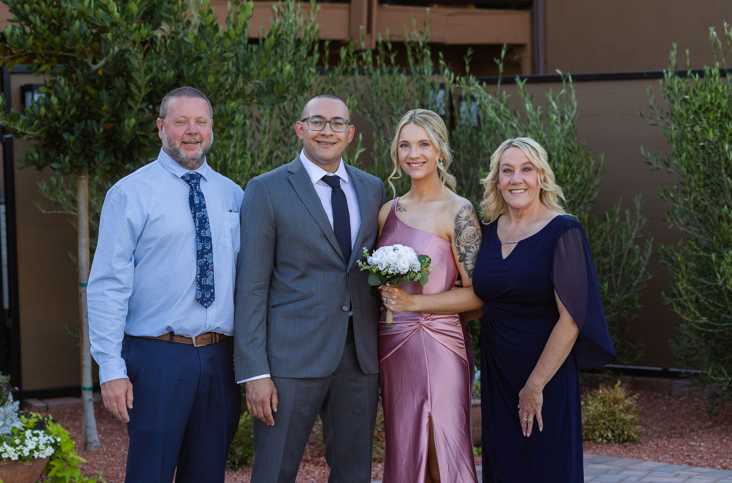 Family group photo at a wedding with four adults posing outdoors, woman in pink bridesmaid dress holding bouquet.