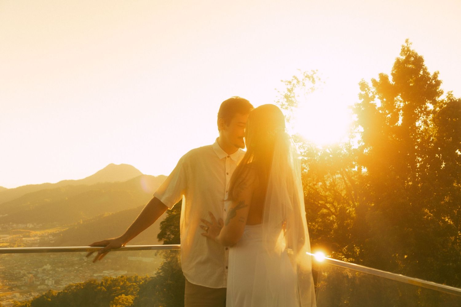 Bride and groom share romantic sunset moment on balcony with golden mountain backdrop on wedding day.
