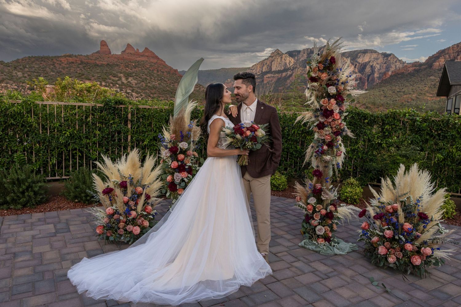 Wedding couple poses in a garden setting with floral arrangements and pampas grass decorations at sunset.