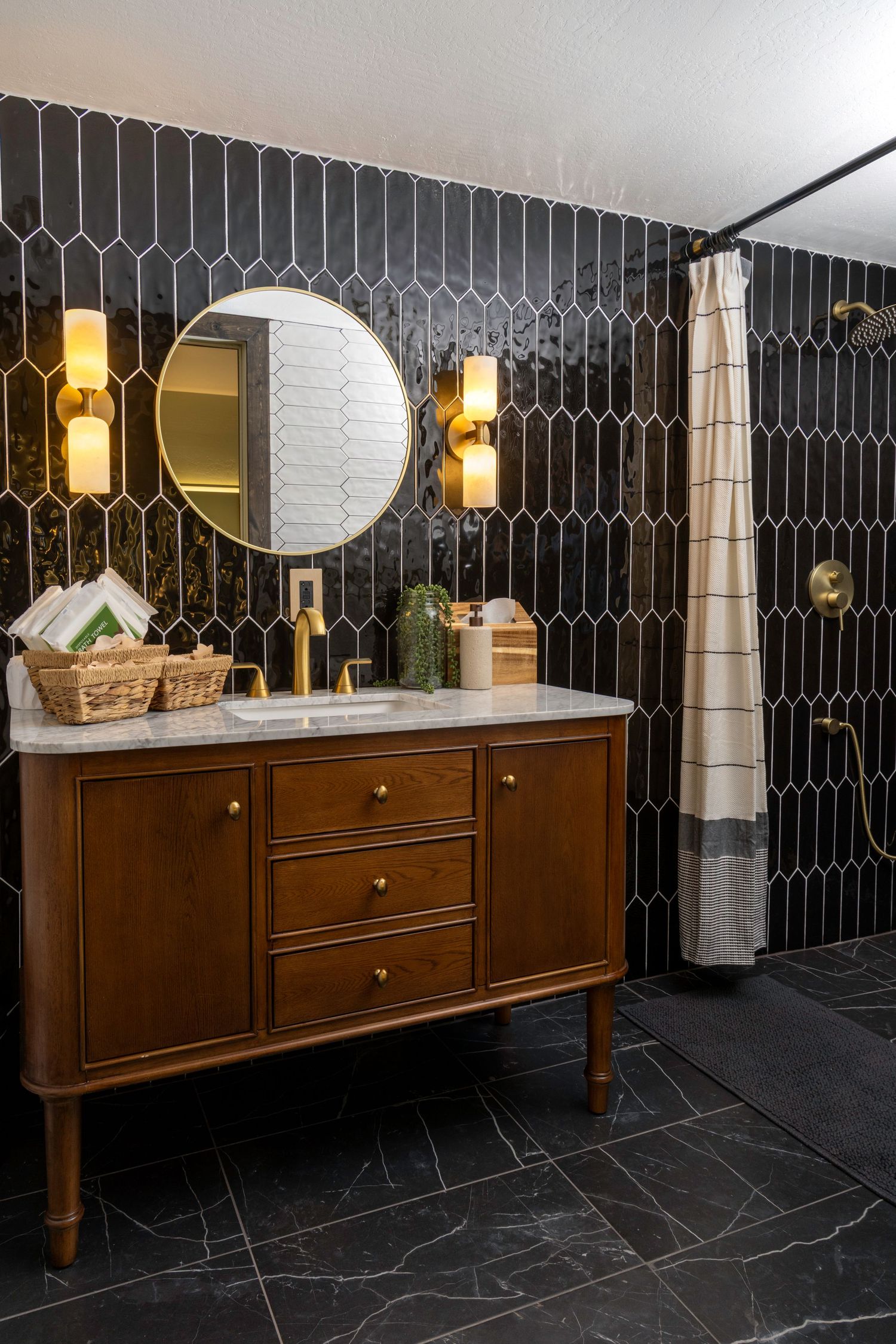 Elegant bathroom with black picket tile walls, wood vanity, gold fixtures, round mirror, and striped shower curtain.