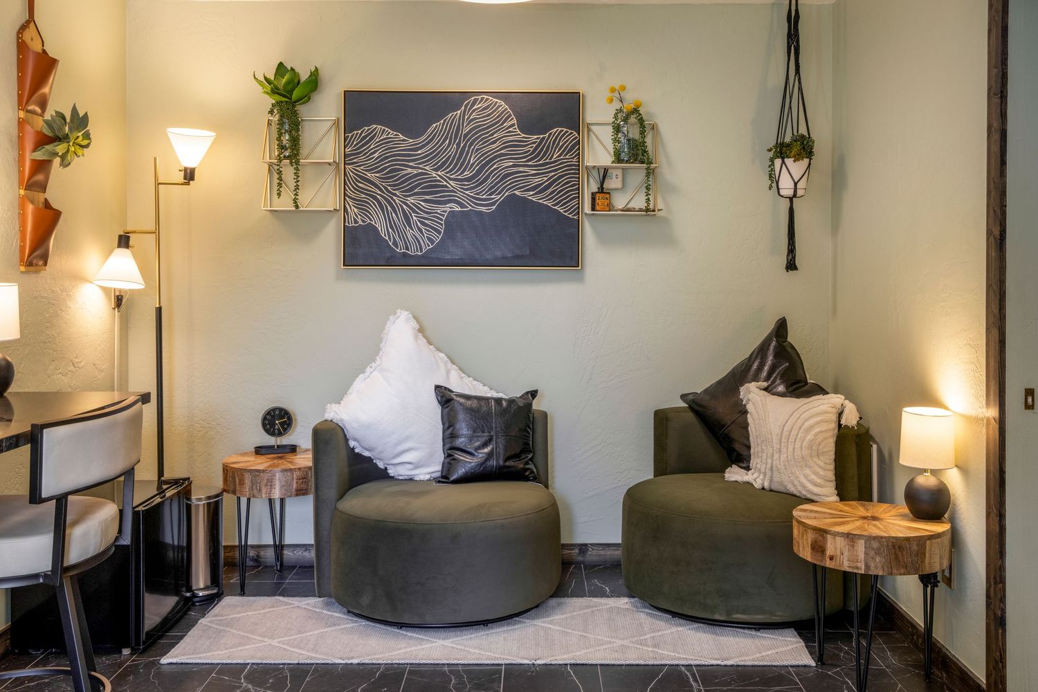 Cozy modern sitting area with two olive green chairs, decorative wall art, shelving with plants, and warm ambient lighting.