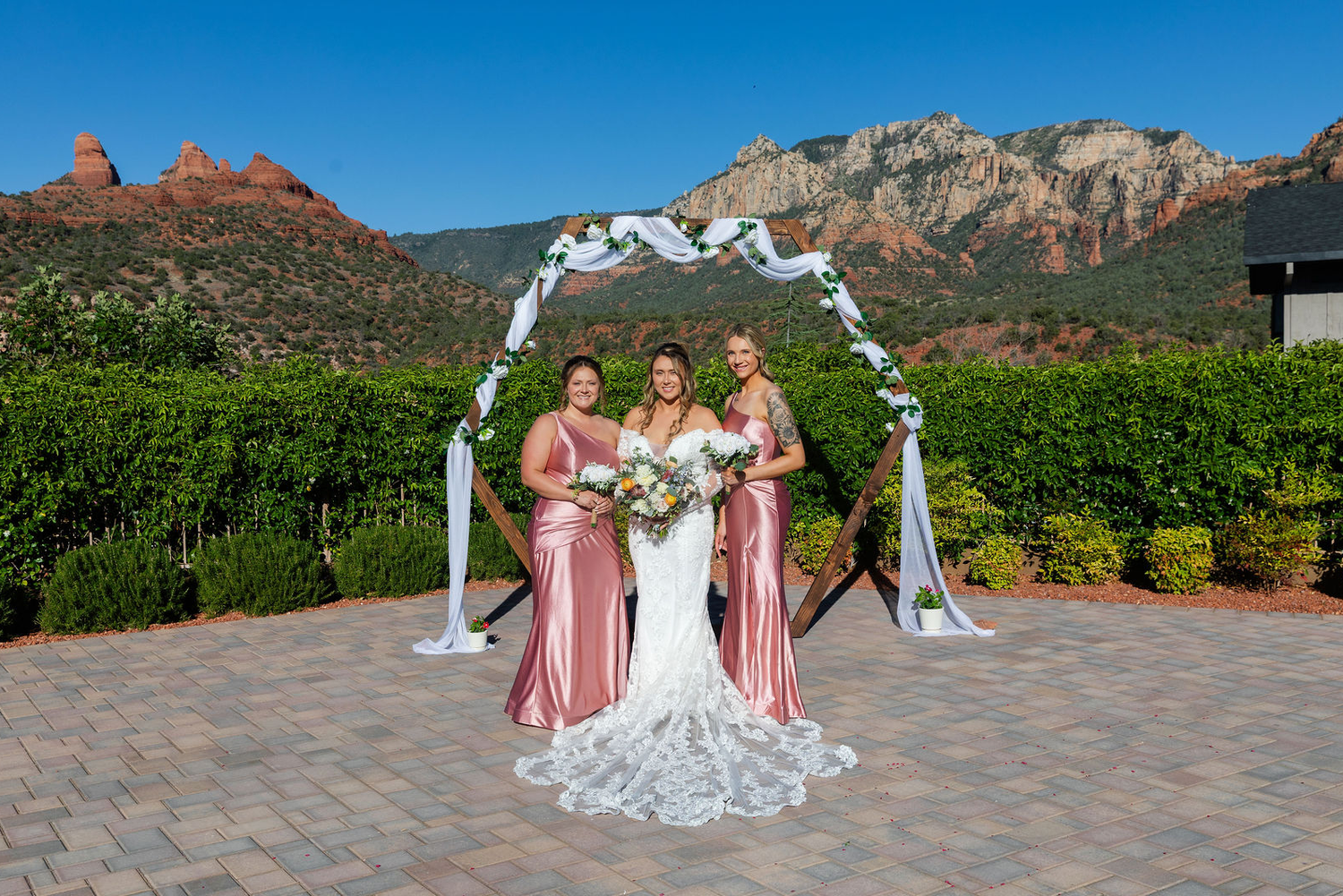 Bride and two bridesmaids in pink dresses pose under floral arch with Sedona red rock mountains backdrop.