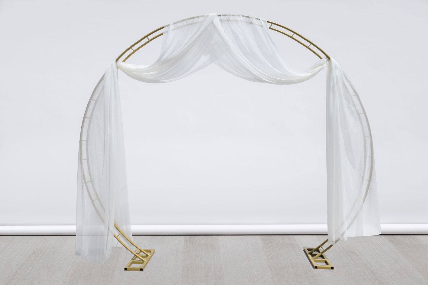 Round gold wedding arch with symmetrical white fabric draping against clean white wall.