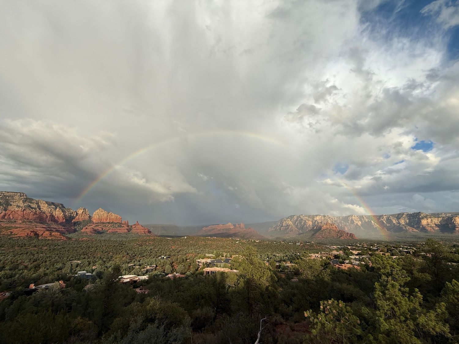 A stunning rainbow arches over Sedona's red rock formations beneath dramatic storm clouds and lush green desert landscape.