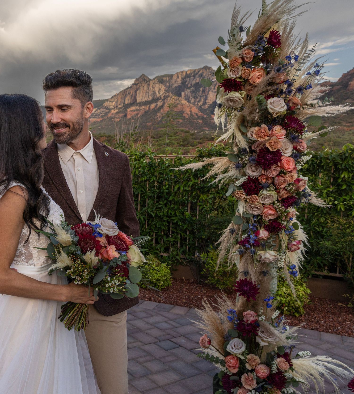 A romantic wedding scene with floral arrangements against dramatic mountain landscapes and cloudy skies at sunset.