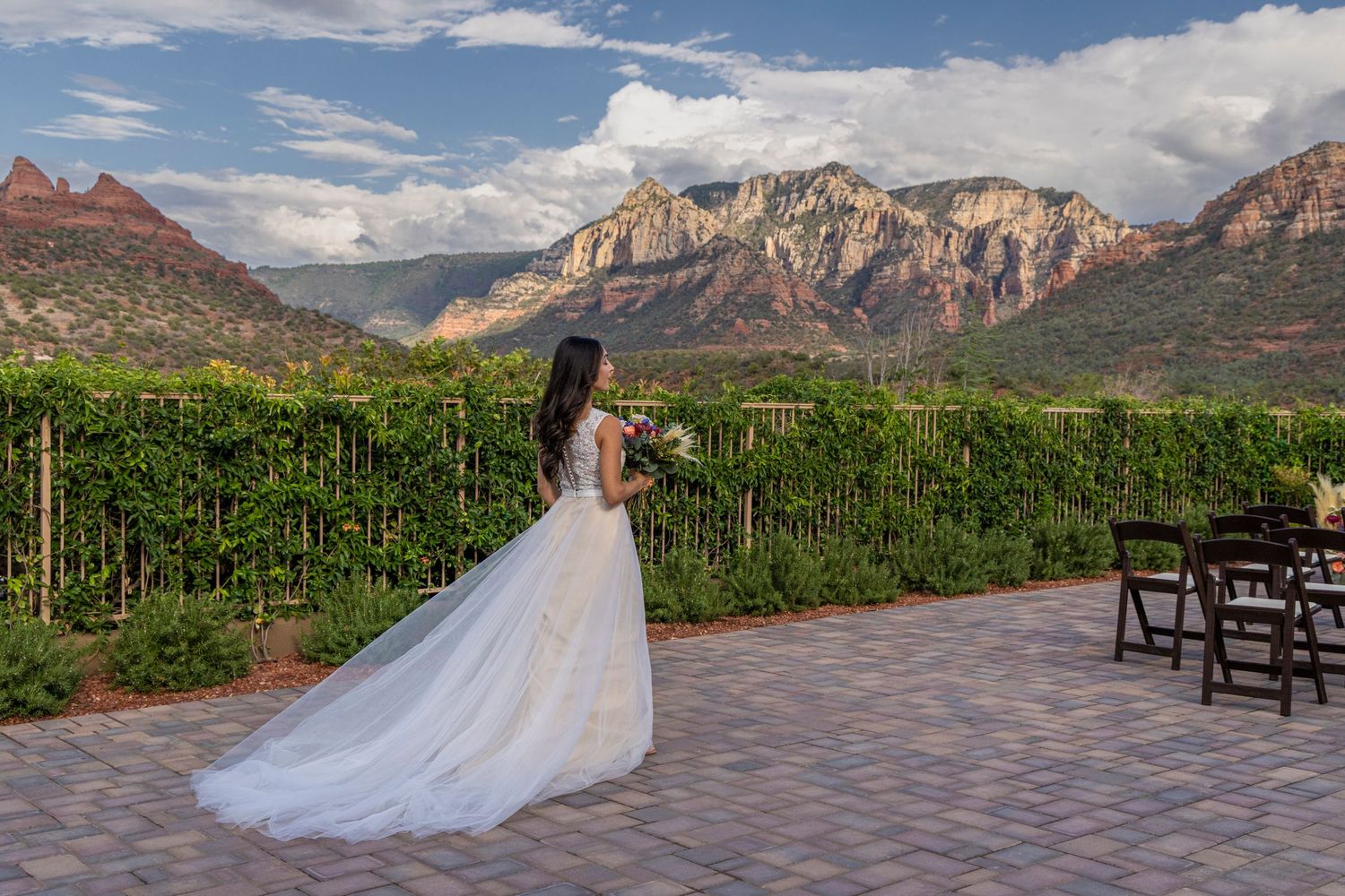 Bride in white gown with long train stands on patio overlooking lush vineyard and red rock mountains in Sedona, AZ.