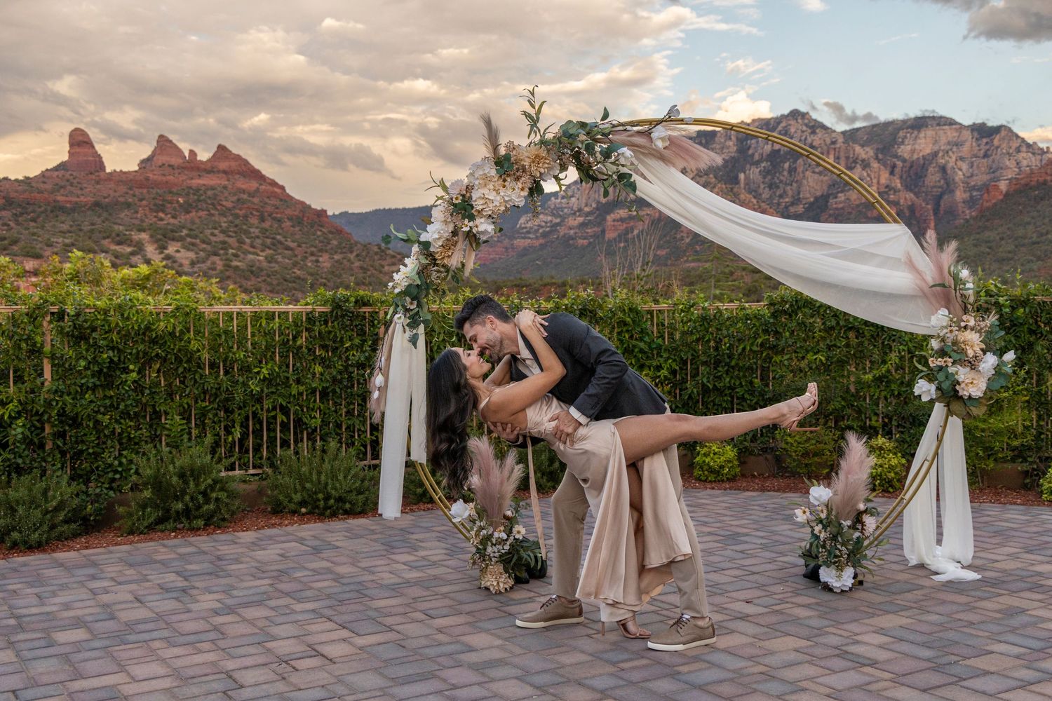 Couple shares romantic dip kiss under floral arch at Sedona wedding venue with red rock mountains at sunset.
