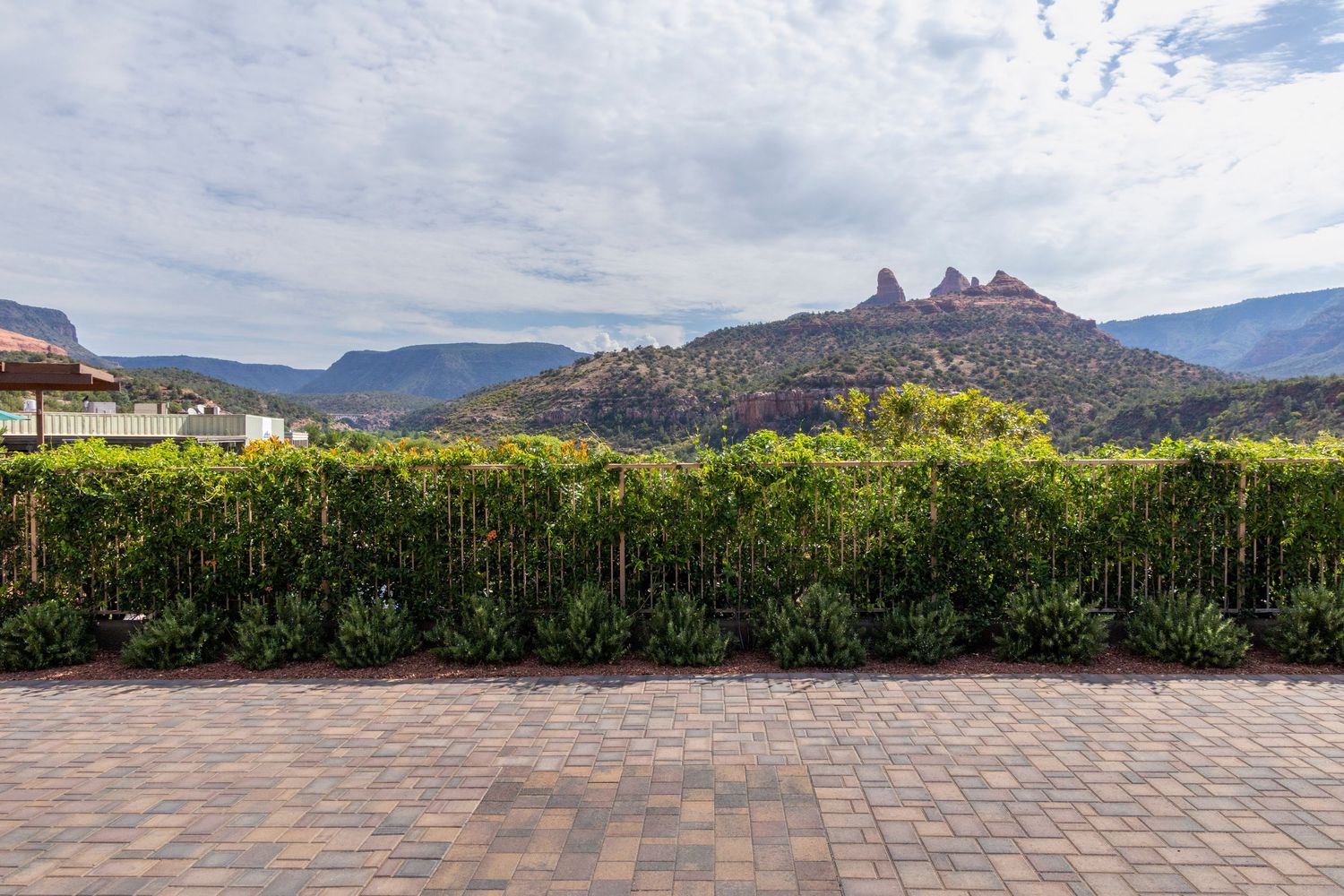 Brick patio with lush green hedge and stunning red rock mountain views under a cloudy sky in Sedona, Arizona.