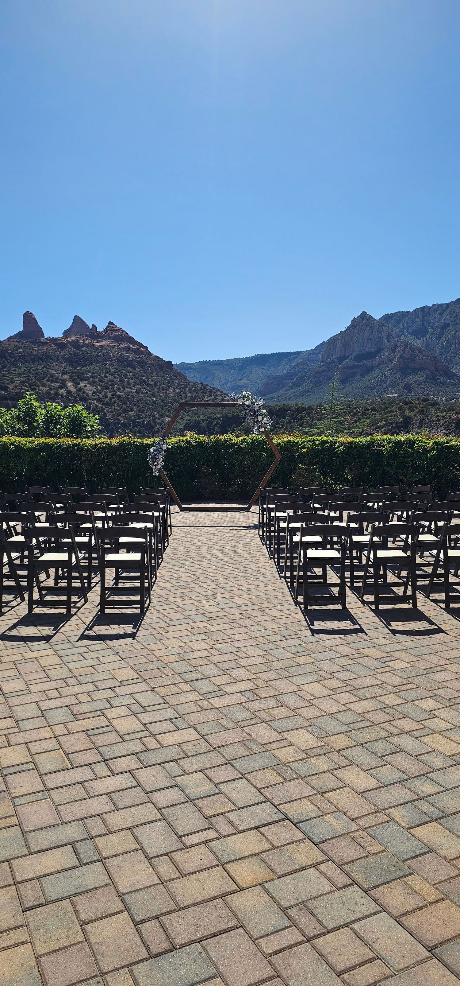 Outdoor wedding ceremony setup with dark wooden chairs, floral arch, and stunning red rock mountain backdrop under clear blue sky.