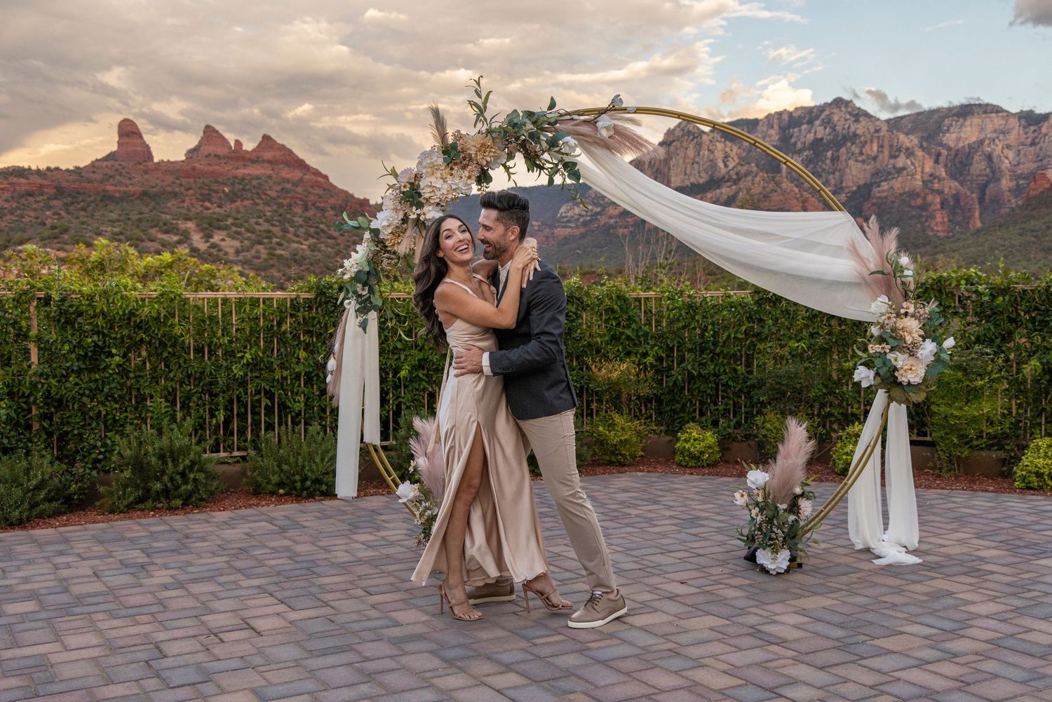 A romantic outdoor wedding ceremony at sunset with red rock mountains in the background and flowing white drapery decorations.