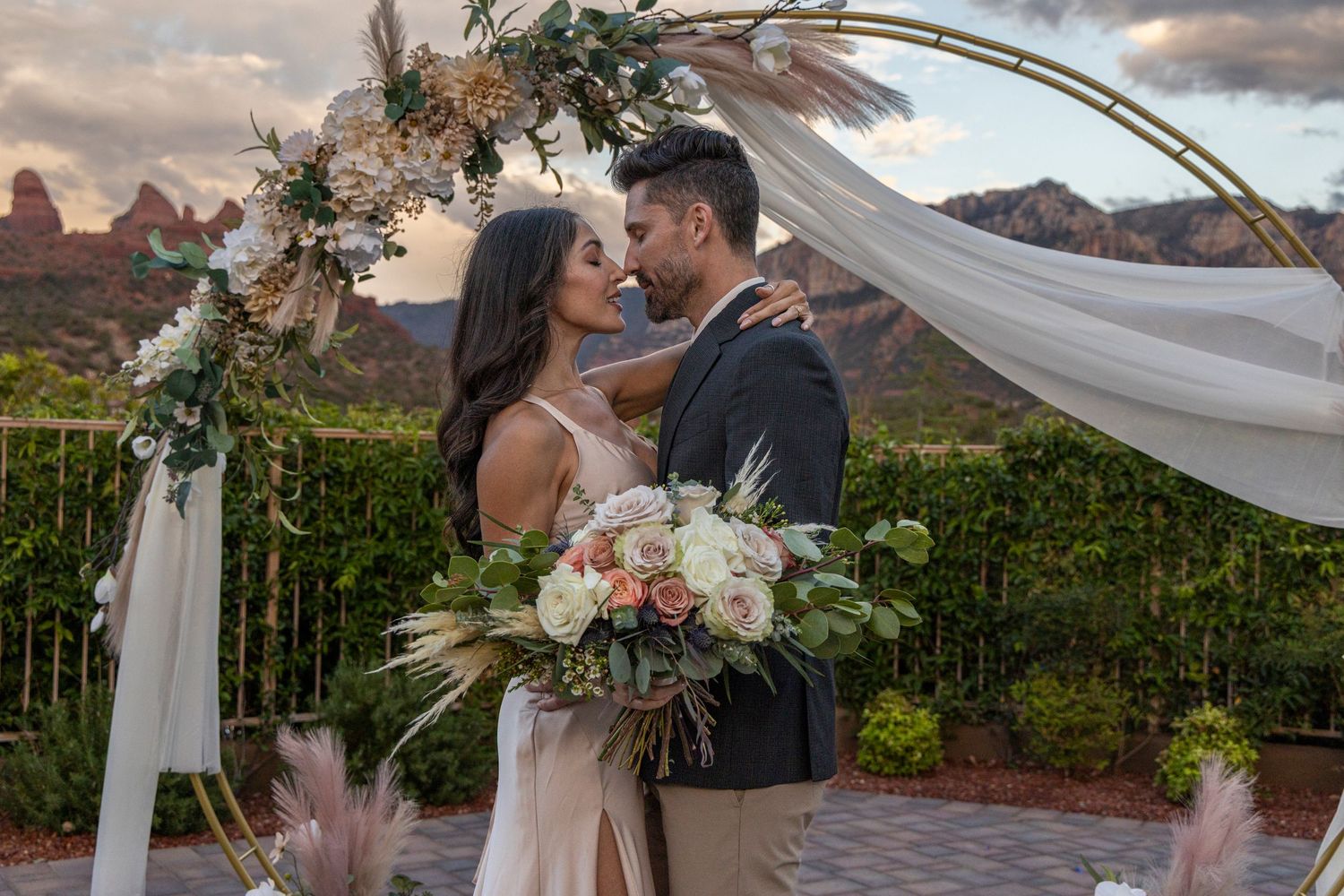 Wedding ceremony setup with floral arch and flowing white fabric against dramatic mountain backdrop at sunset.