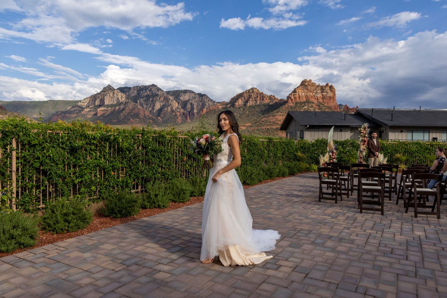 A scenic outdoor wedding venue with mountain backdrop and stone patio surrounded by lush greenery and dark wood chairs.