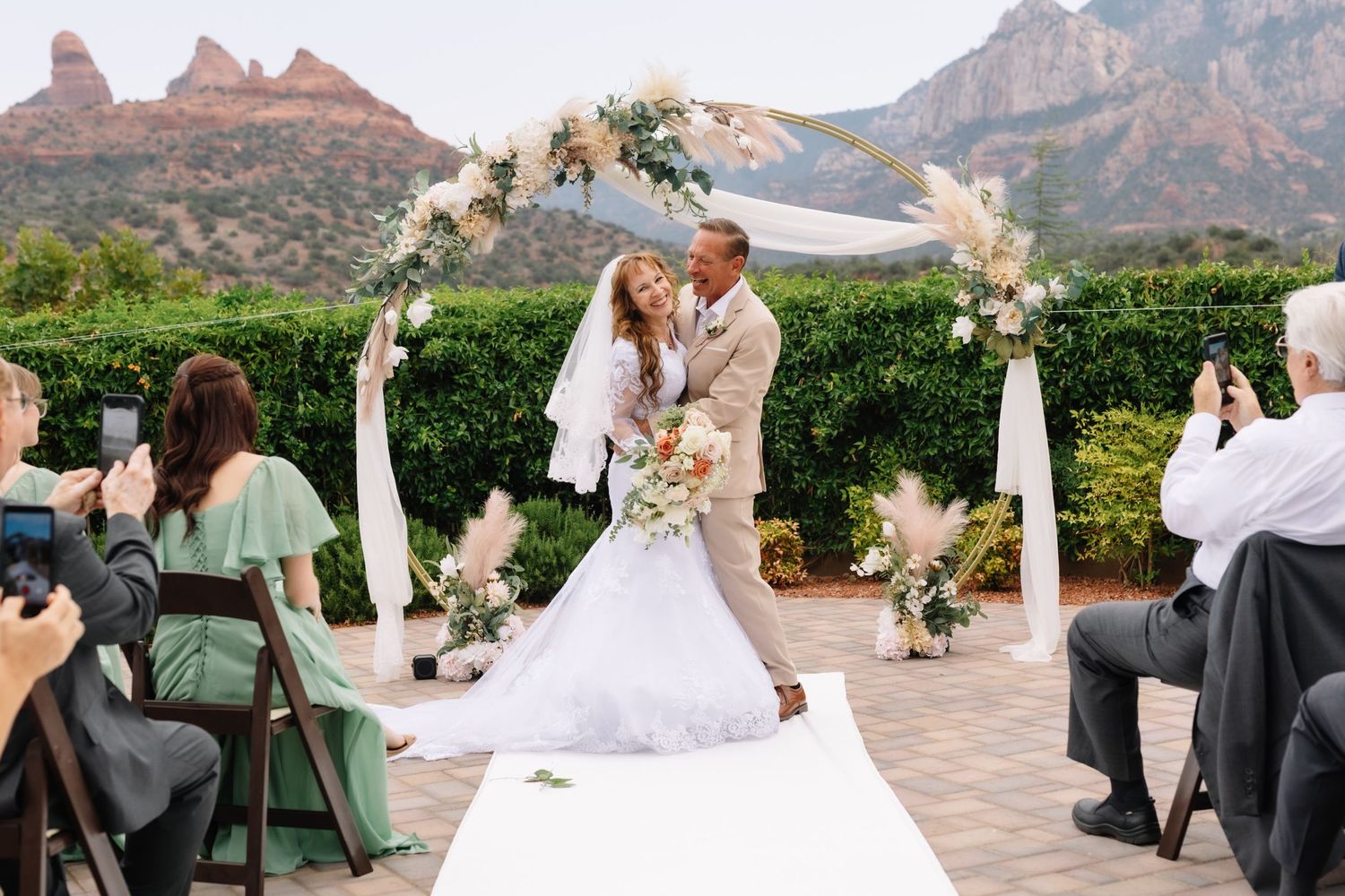 Bride and groom share first kiss under floral arch with stunning red rock Sedona mountains backdrop at outdoor wedding.