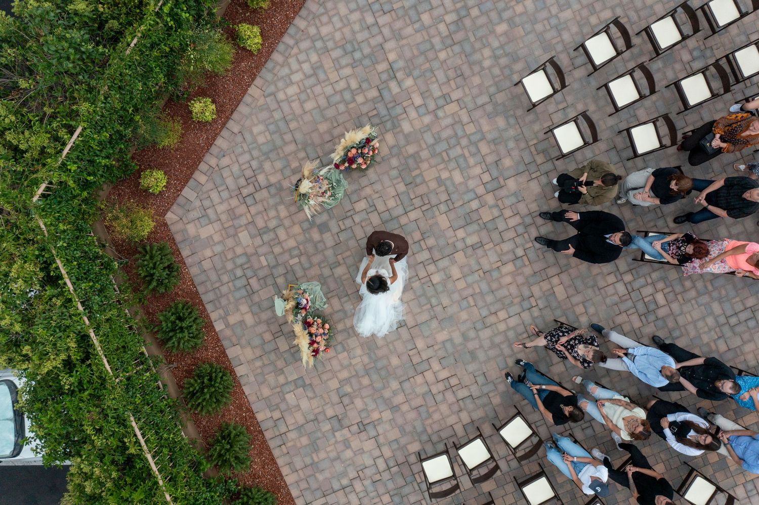 Aerial view of people sitting in white chairs arranged in rows on a brick patio surrounded by greenery.