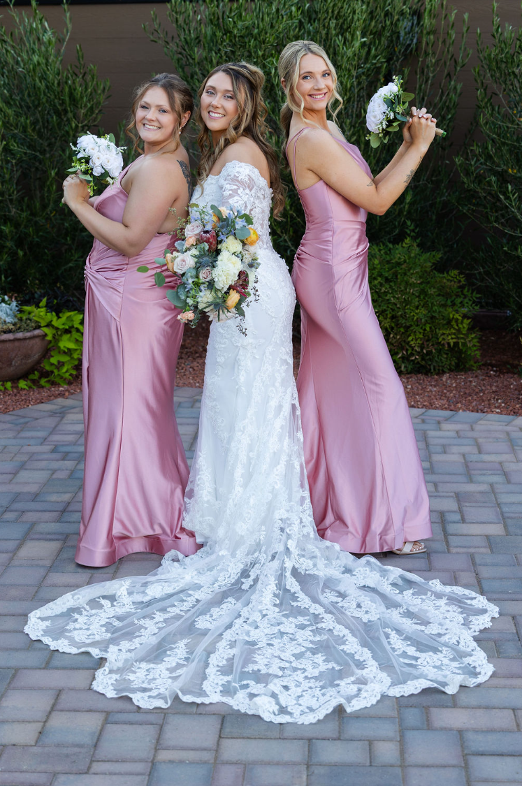 Bride in lace gown with cathedral train poses with two bridesmaids in pink satin dresses holding bouquets outdoors.