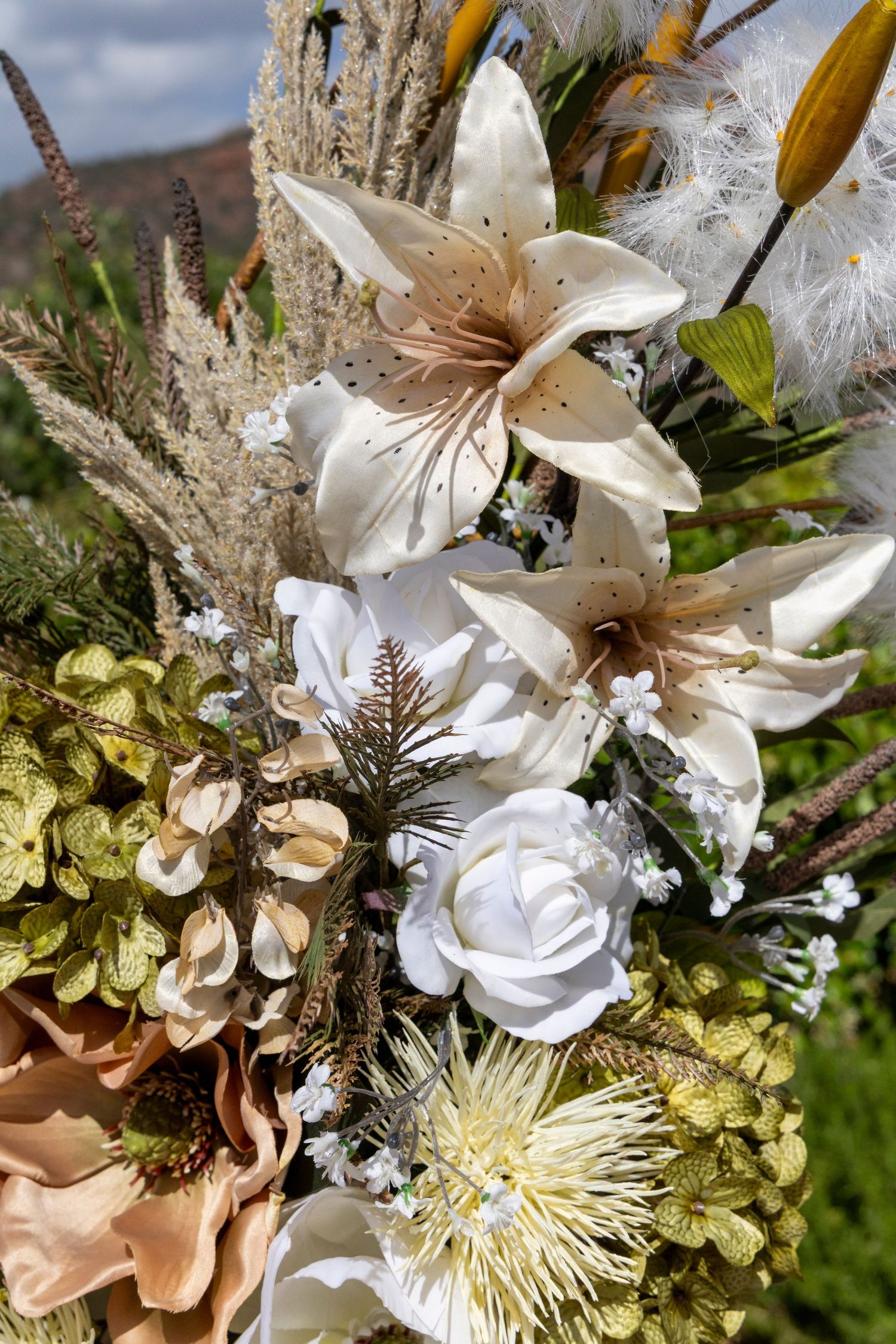 Elegant floral arrangement featuring white lilies, roses, and dried botanicals in neutral tones against a natural outdoor backdrop.
