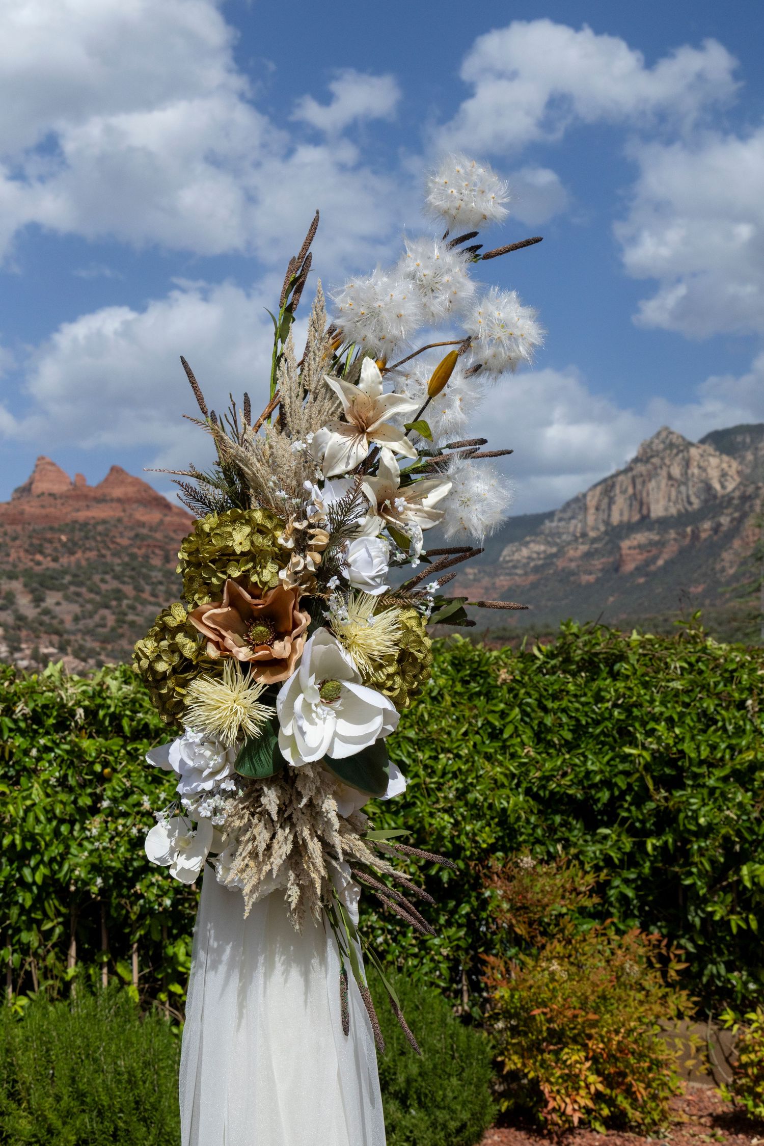 Elegant floral arrangement with white blooms and dried grasses against Sedona red rock mountains and blue sky backdrop.