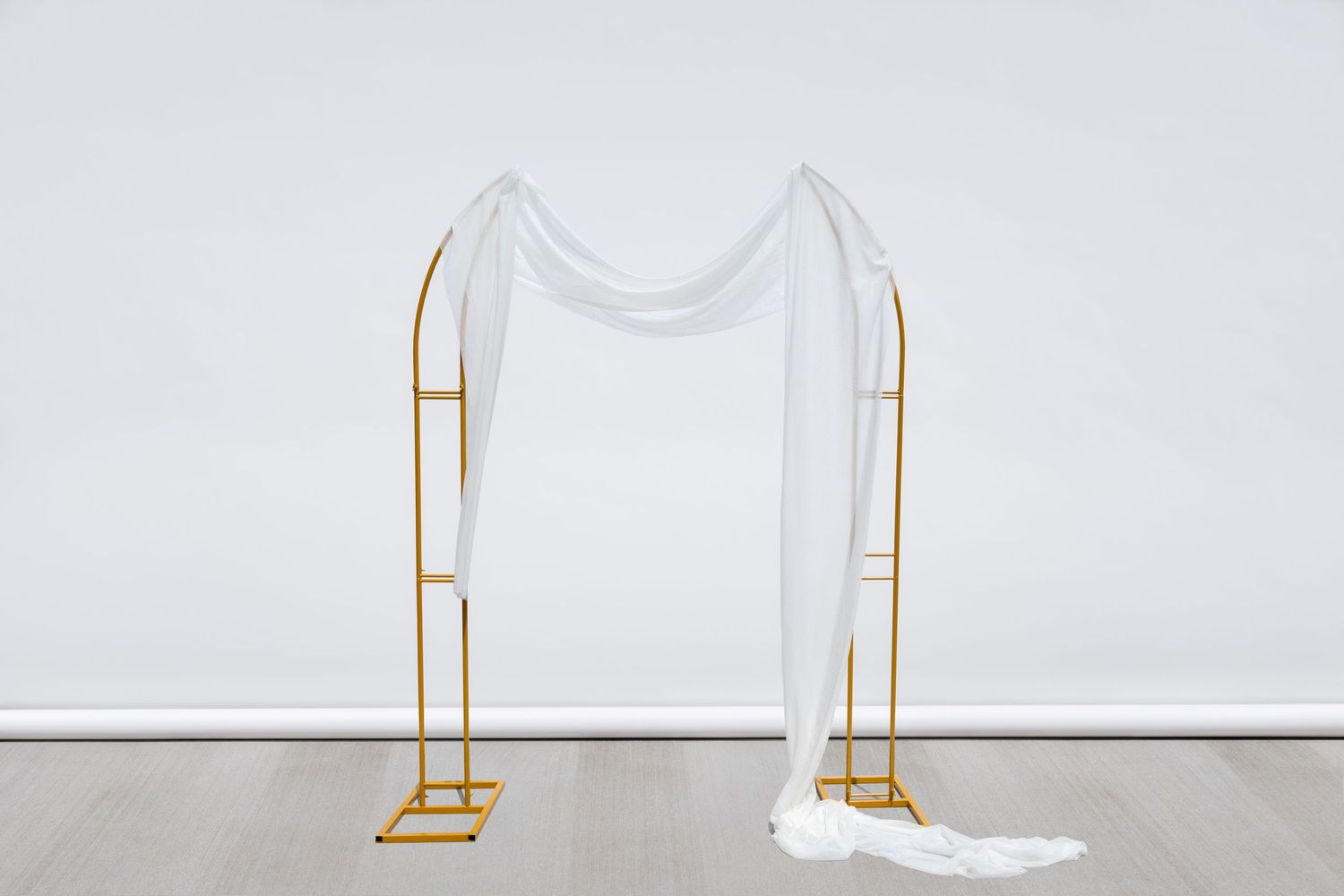 Gold metal arch frame with asymmetrical white fabric draping against white background.