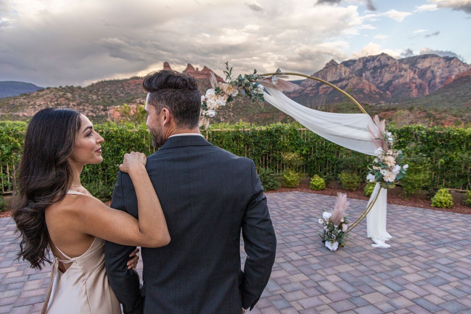 A couple stands on a patio overlooking desert mountains with a white fabric arch and floral arrangements in view.