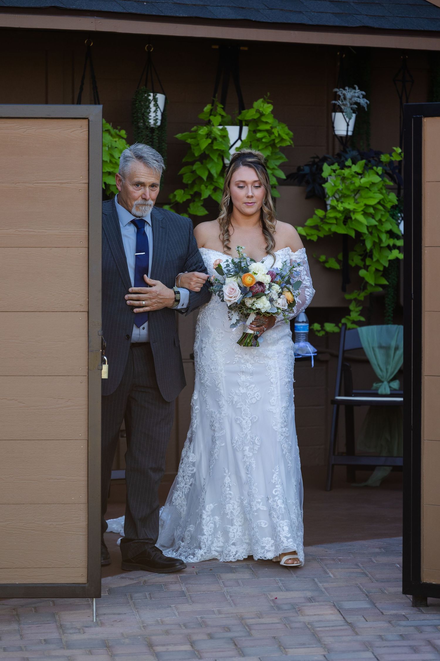 Bride in lace gown holding bouquet walks with older man in suit through decorated venue entrance.