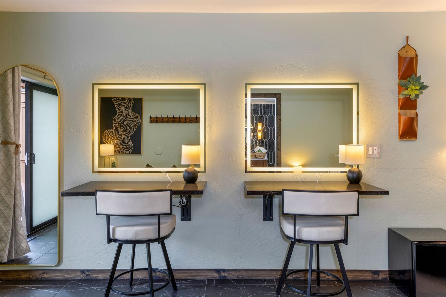 Two illuminated vanity mirrors with white bar stools at a dual makeup station in a modern hotel room.