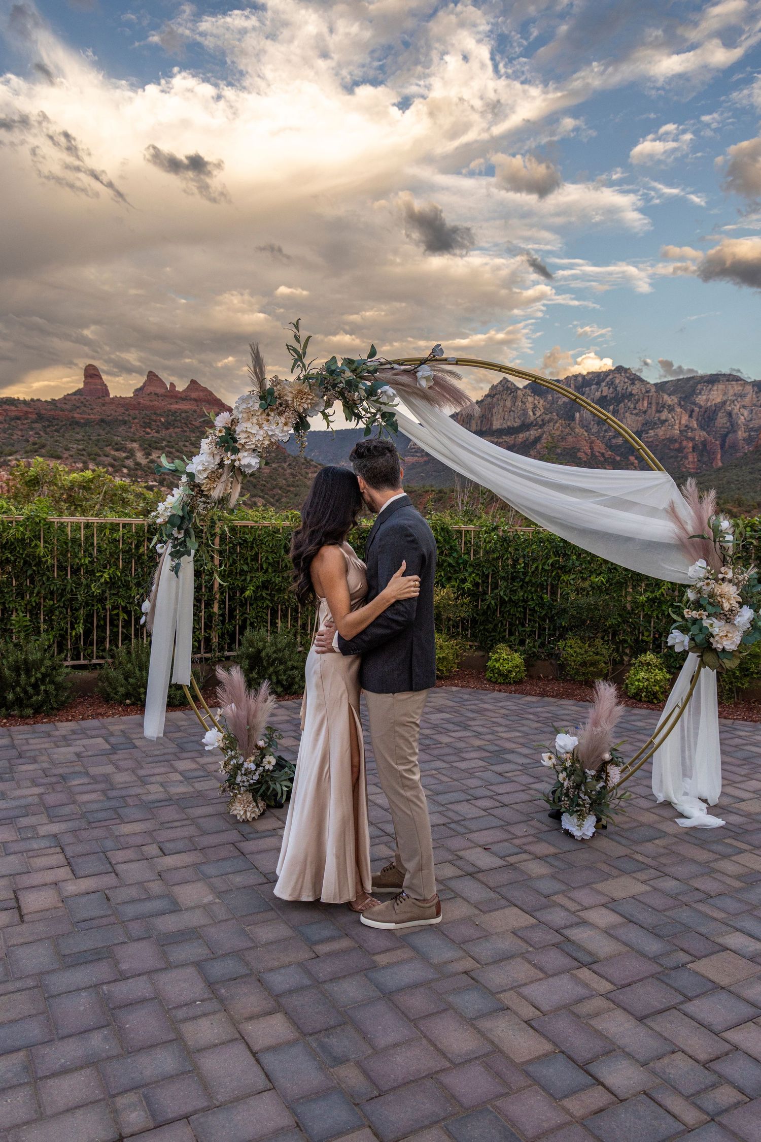 Couple embracing under floral arch at sunset wedding in Sedona, Arizona with red rock formations in background.