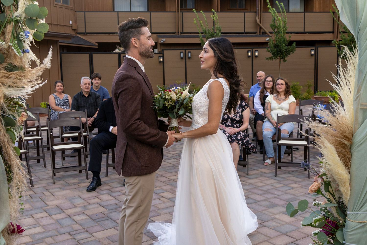 A couple shares a romantic first dance at an outdoor wedding reception surrounded by guests and pampas grass decor.