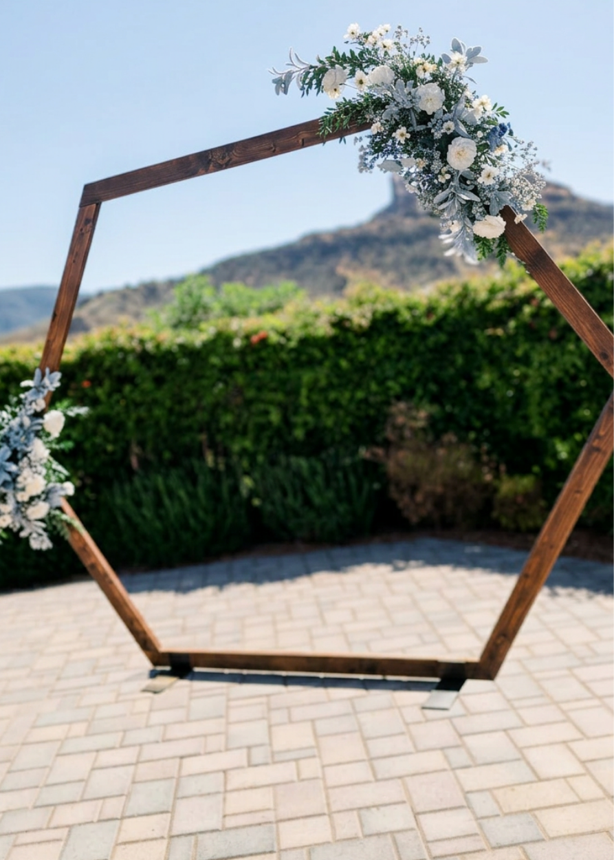 Rustic hexagonal wooden wedding arch adorned with white roses and greenery, set against a scenic mountain backdrop.