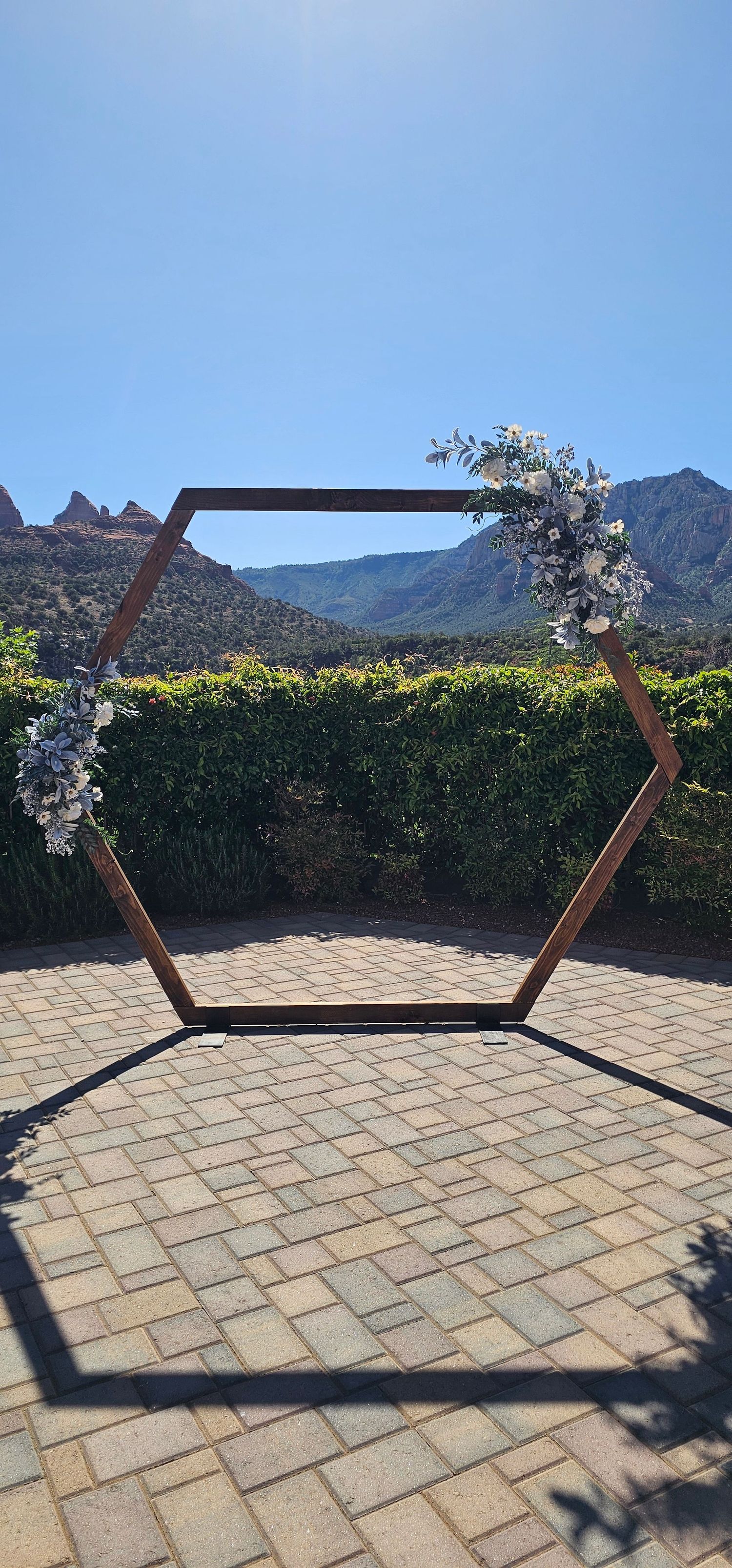 Hexagonal wooden wedding arch with white floral arrangements, set on brick patio with mountain backdrop.