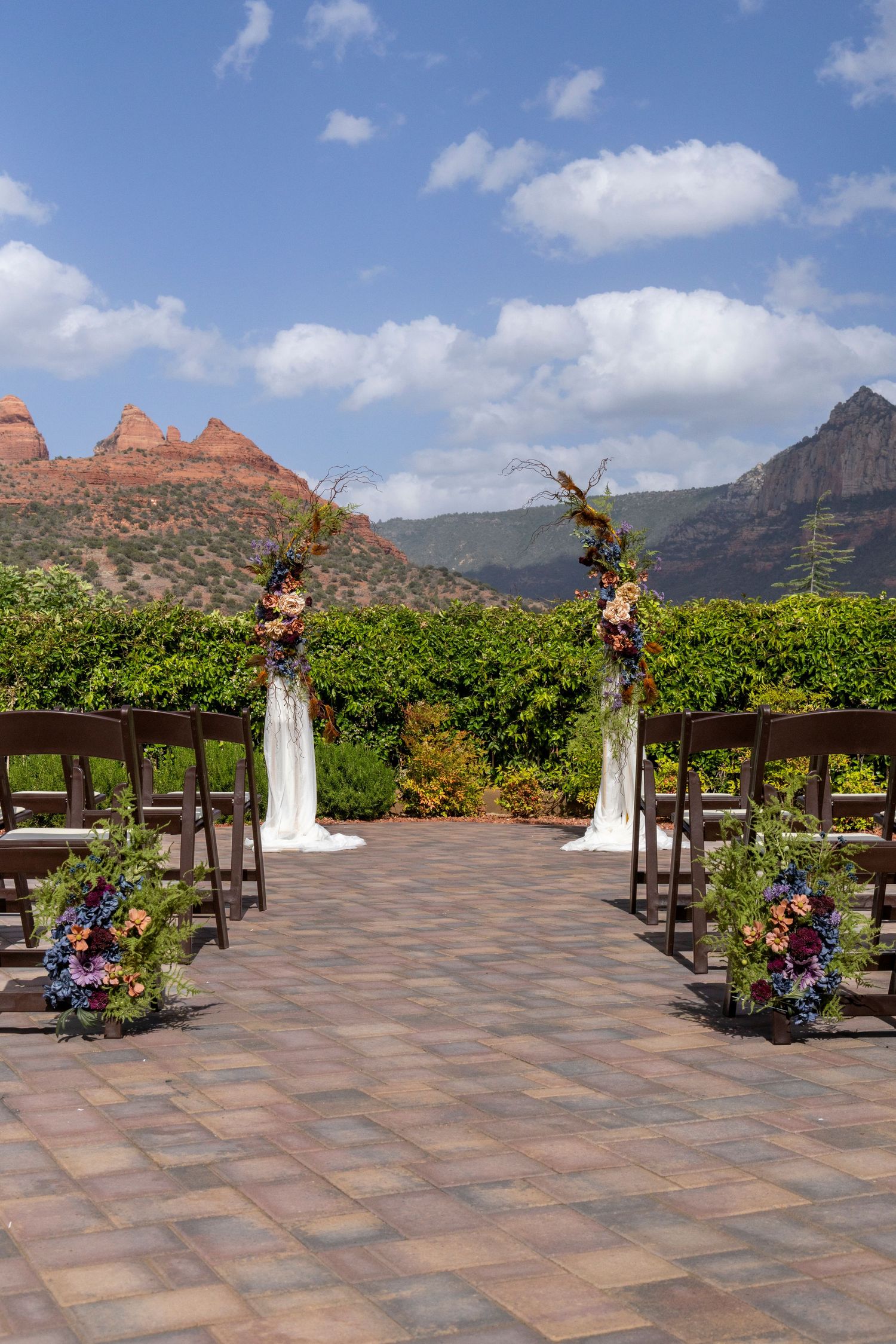 Outdoor wedding ceremony setup with floral arch, wooden chairs, and stunning Sedona red rock mountain backdrop under blue sky.