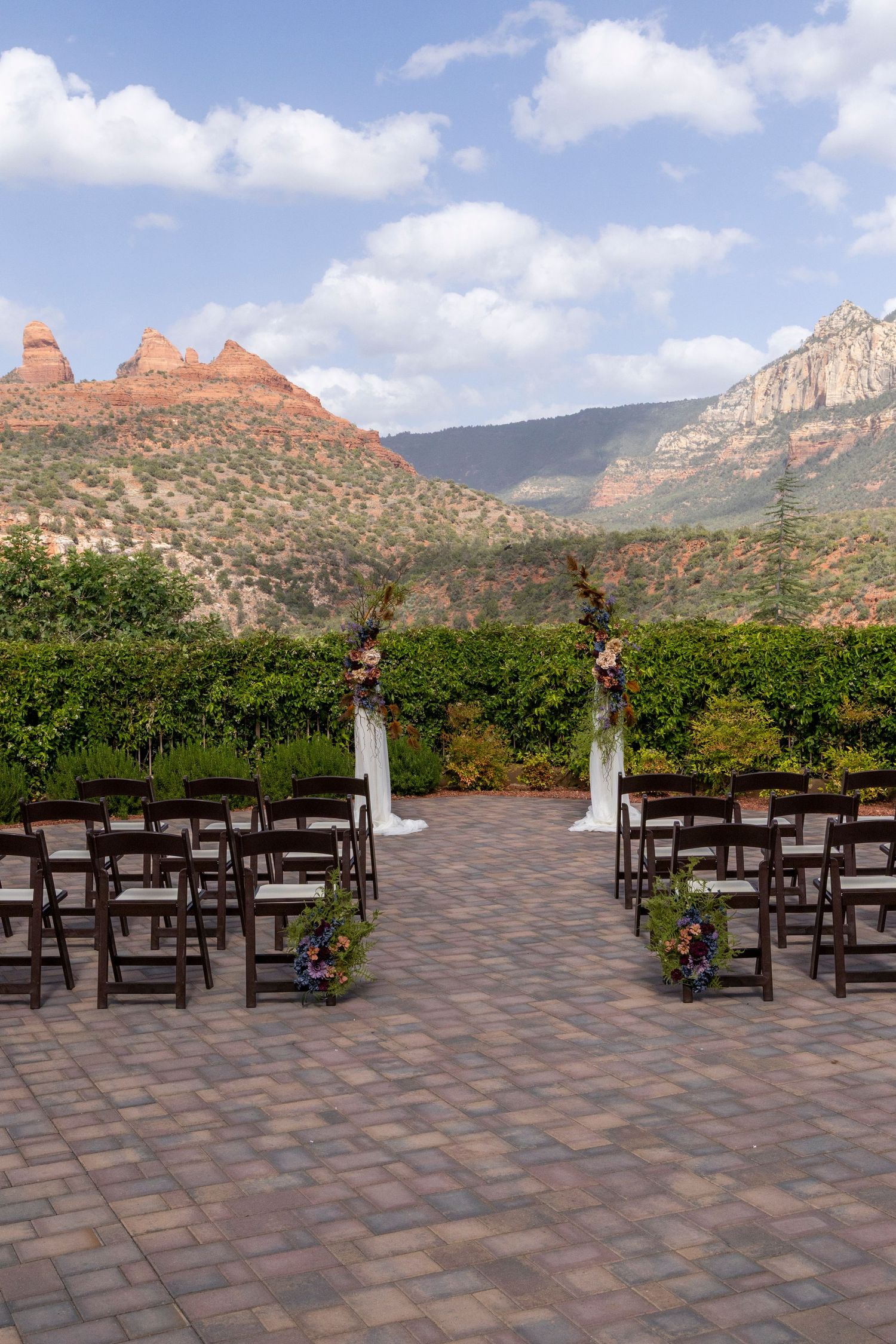 Outdoor wedding ceremony setup with dark wooden chairs on brick patio, floral arch, and stunning Sedona red rock backdrop.