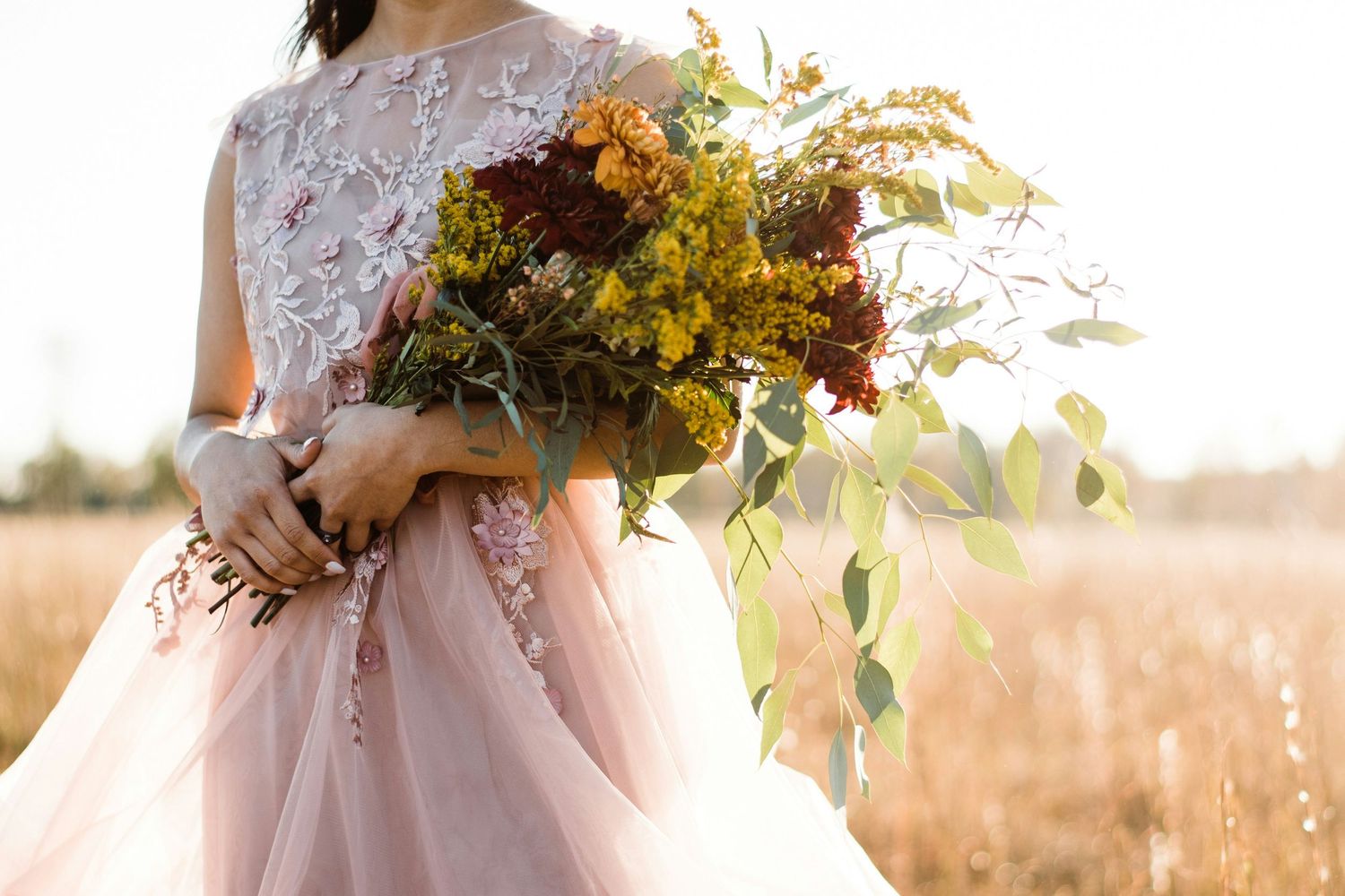 Bride in pink floral gown holding a rustic autumn bouquet with burgundy and yellow wildflowers in a golden field.