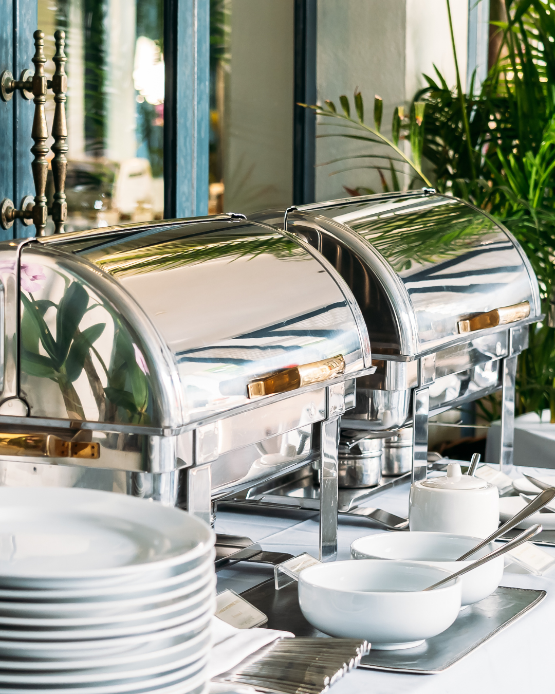 Row of stainless steel chafing dishes and white plates set up for catering service.