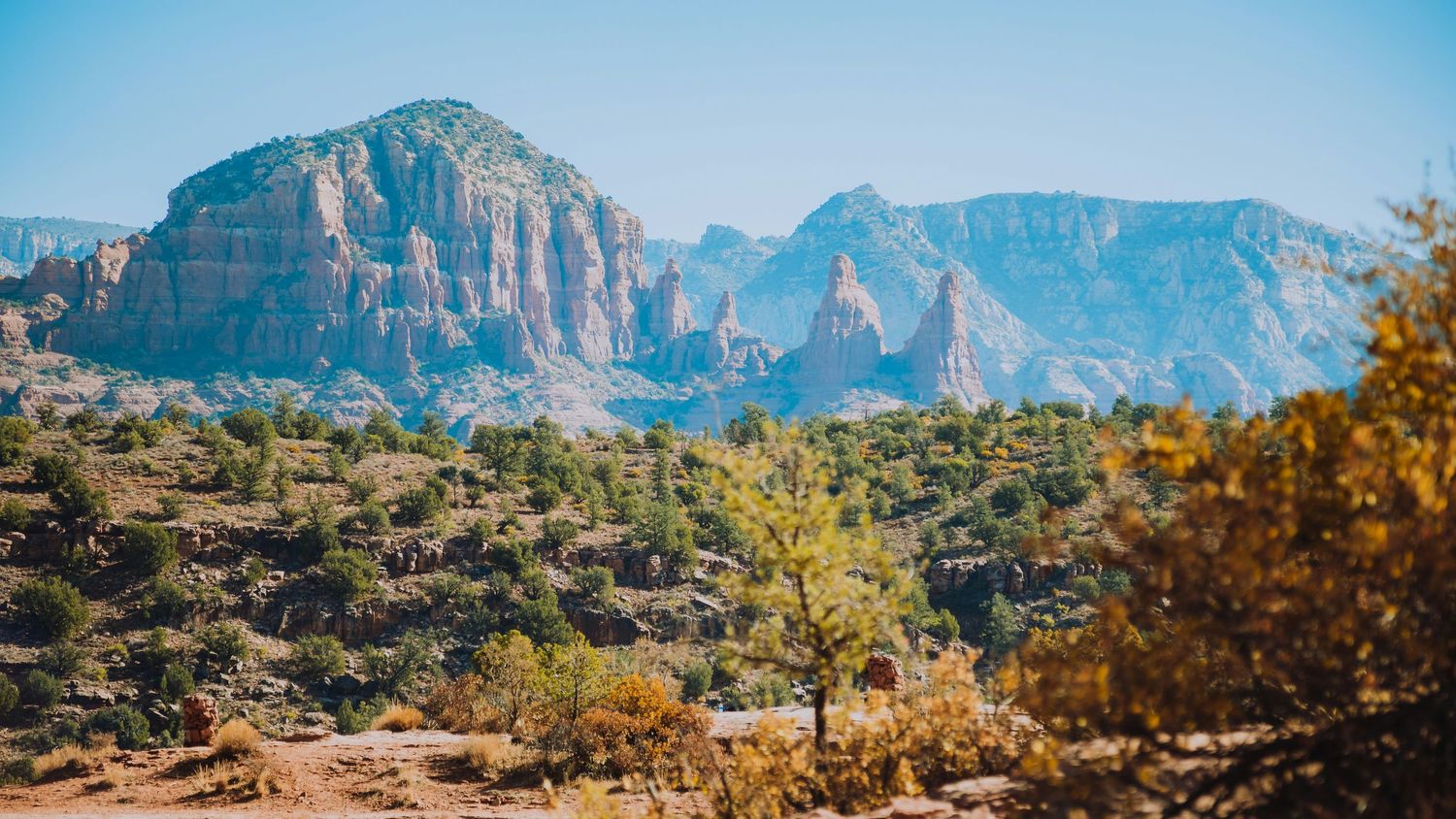 Red rock formations rise above a lush desert landscape with green trees under a clear blue sky in Sedona, Arizona.