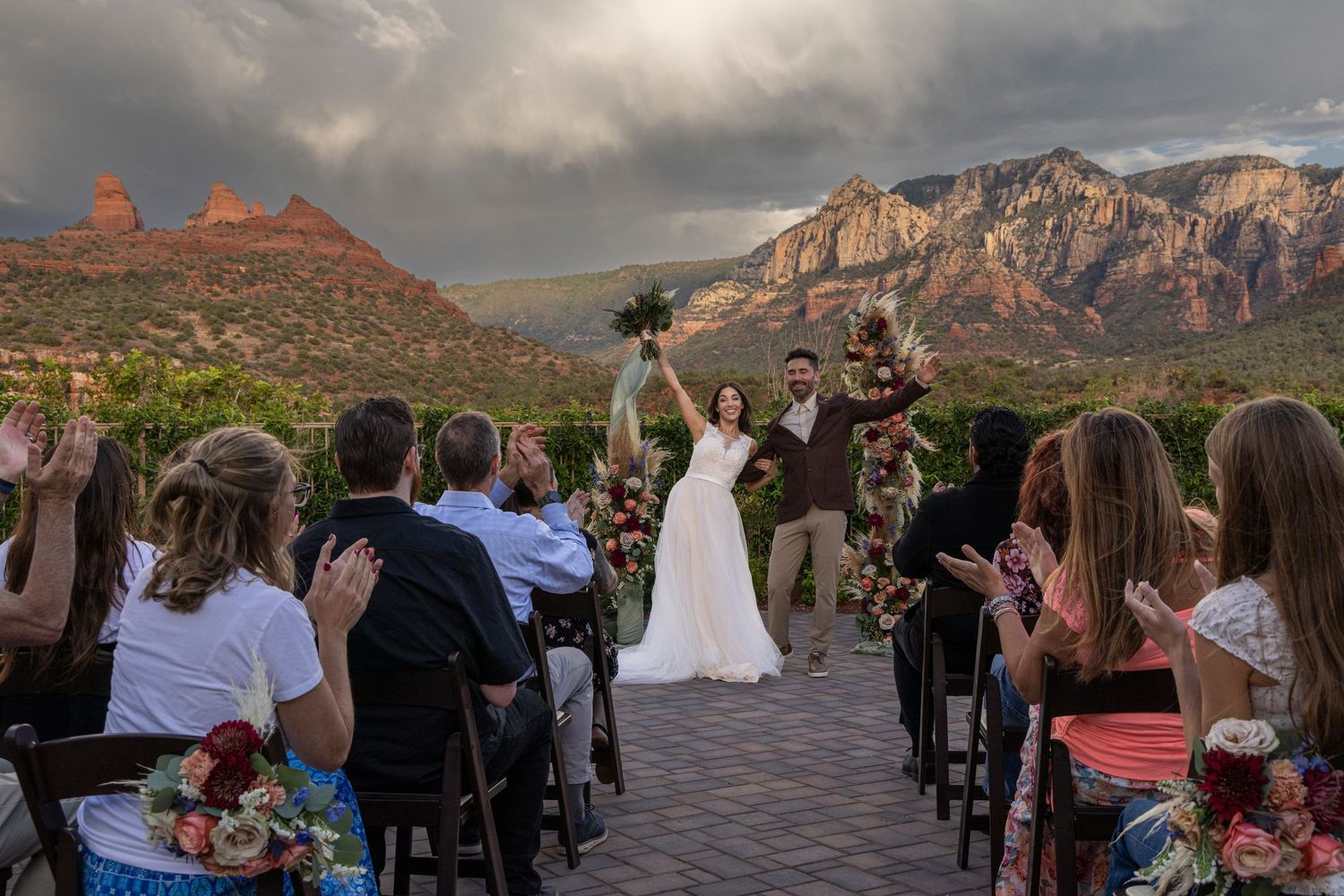 Outdoor wedding ceremony with guests seated facing red rock mountain formations under dramatic cloudy skies.