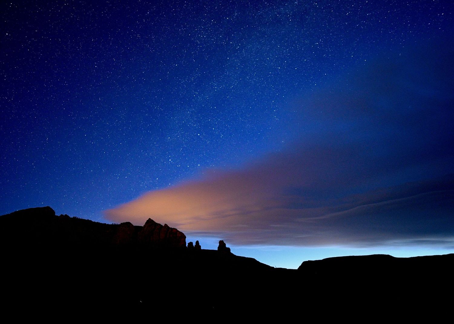 Stunning night sky with stars and glowing clouds over dark rocky silhouettes in a deep blue landscape.