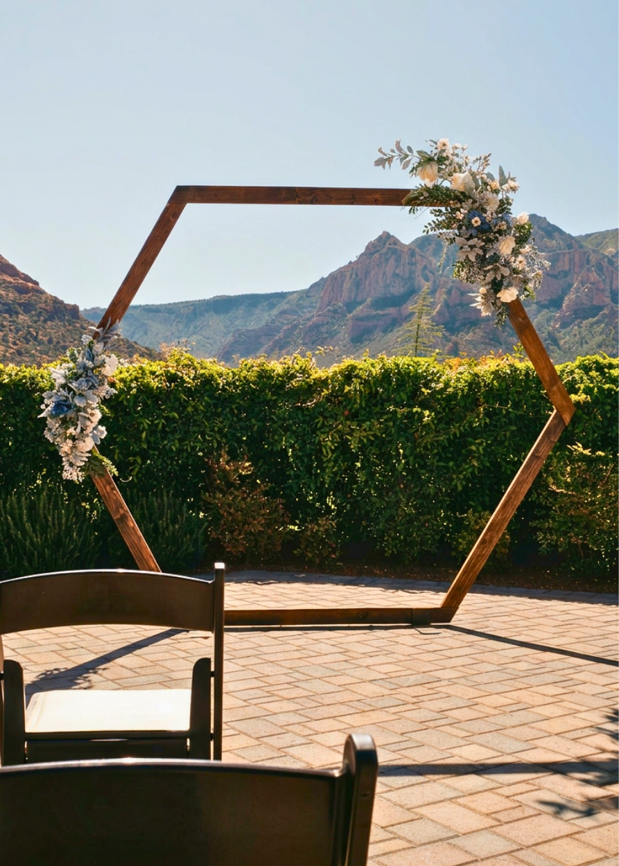 Hexagonal wooden wedding arch adorned with white flowers, set on a brick patio with lush greenery and red rock mountains.