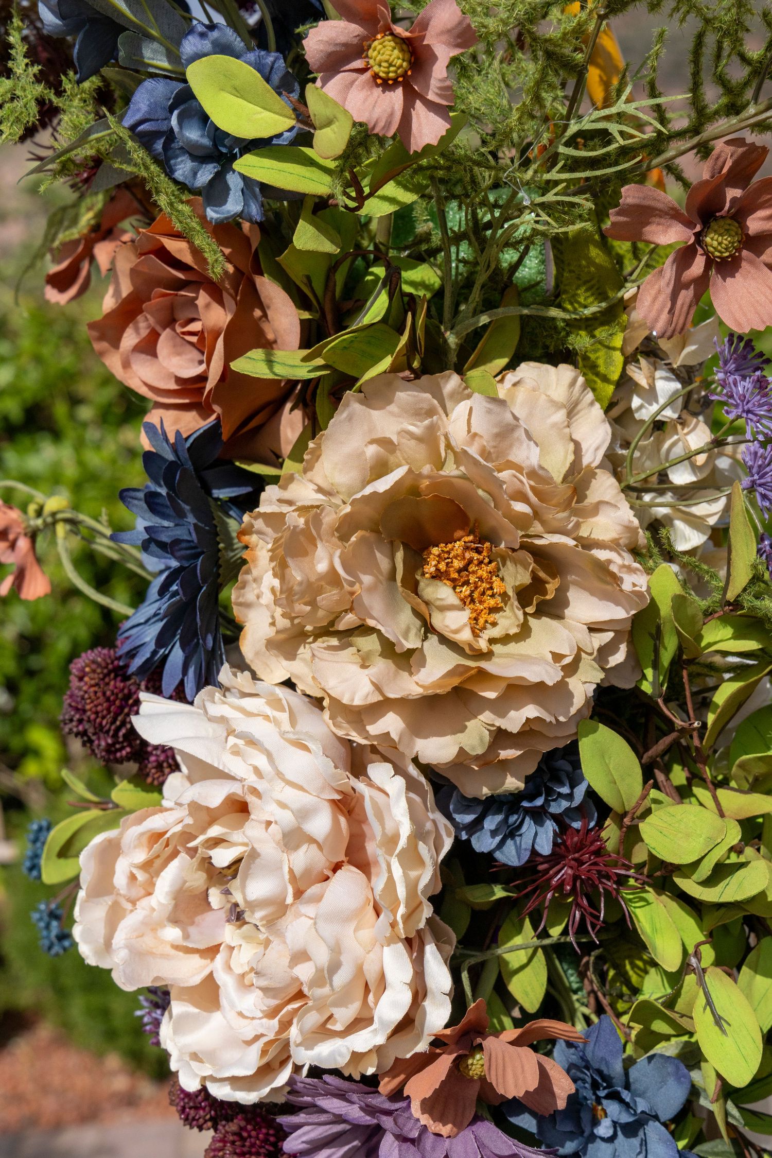 Lush floral arrangement featuring cream peonies, pink roses, blue flowers, and green foliage in a garden setting.