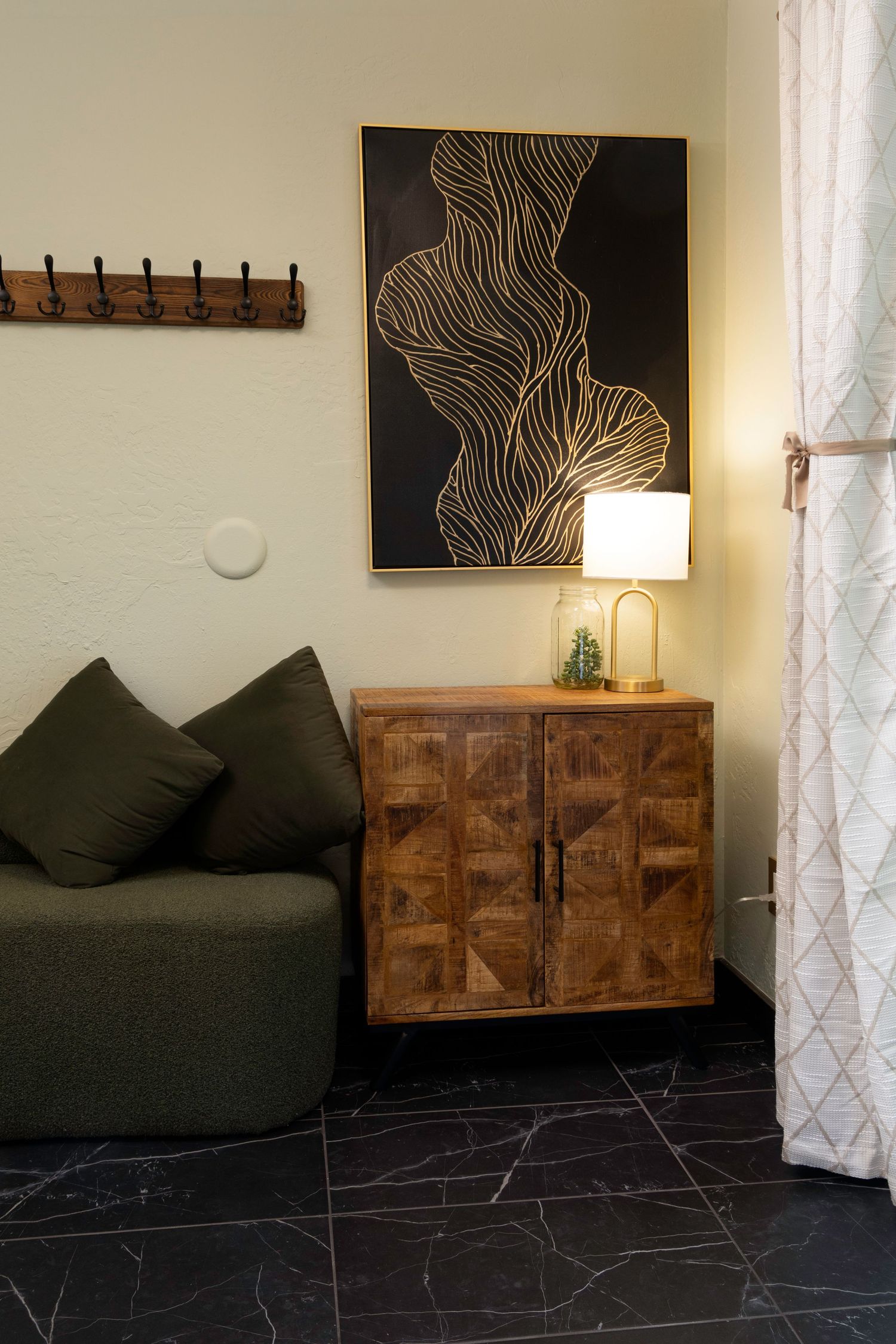 Cozy corner with green sofa, wooden cabinet, gold lamp, abstract black wall art, and marble tile flooring.