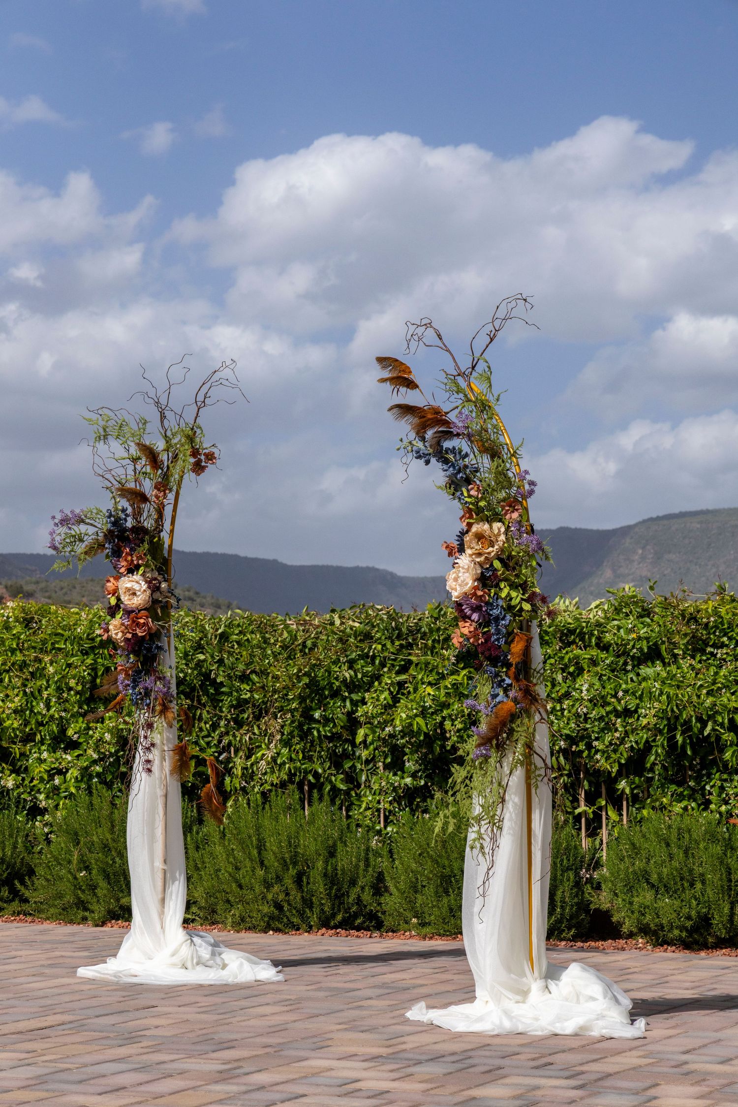 Two tall floral wedding ceremony pillars with white draping, colorful blooms, and greenery backdrop under a partly cloudy sky.